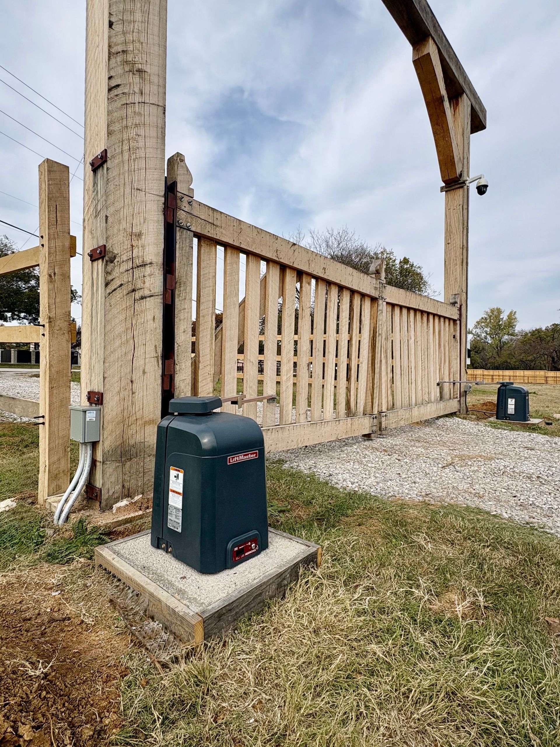 Wooden fence and gate beside a concrete post, with a green utility box on a gravel yard.