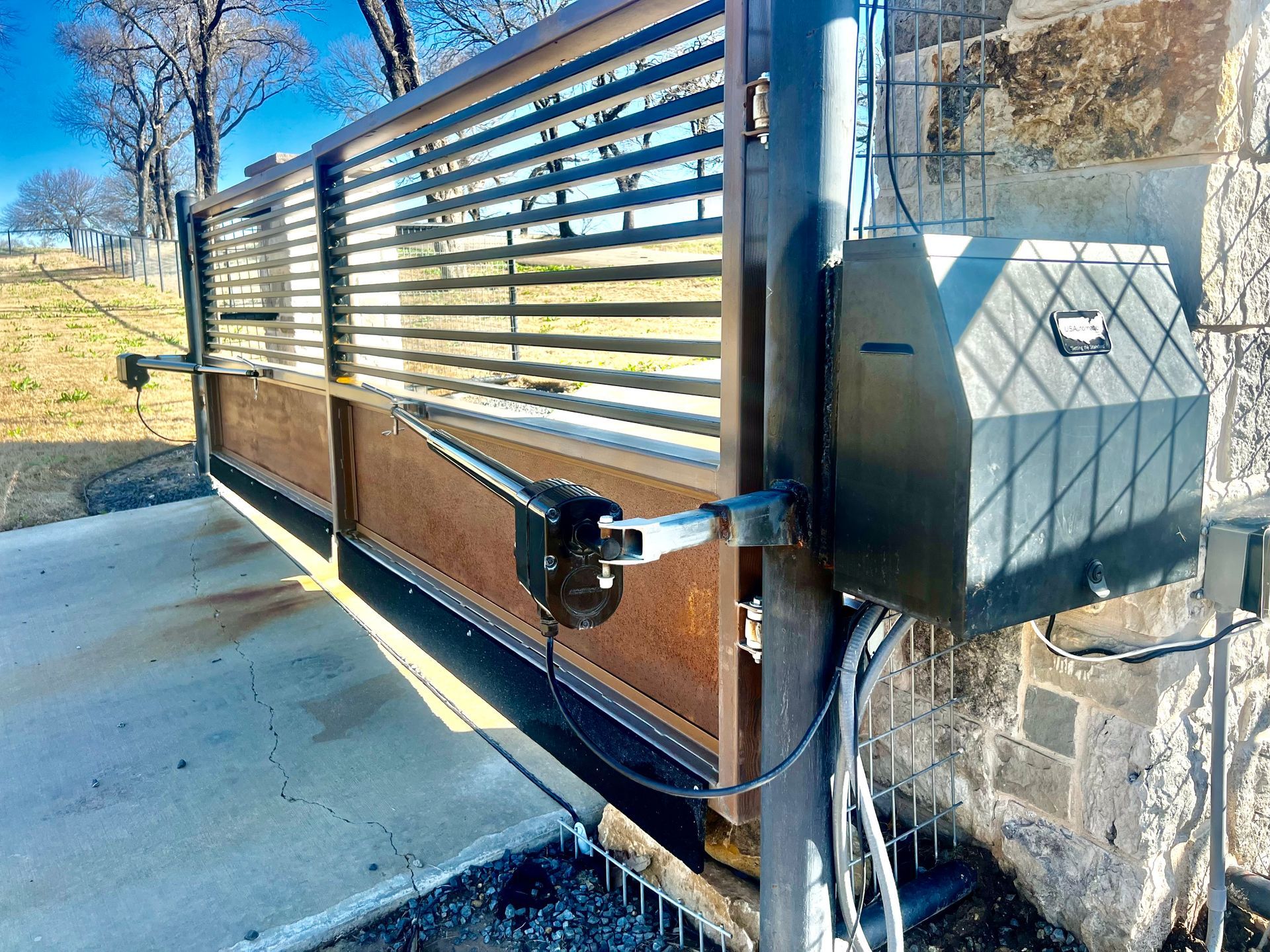 Wooden slat driveway gate with metal post and fence beside a concrete path, viewed in bright sunlight