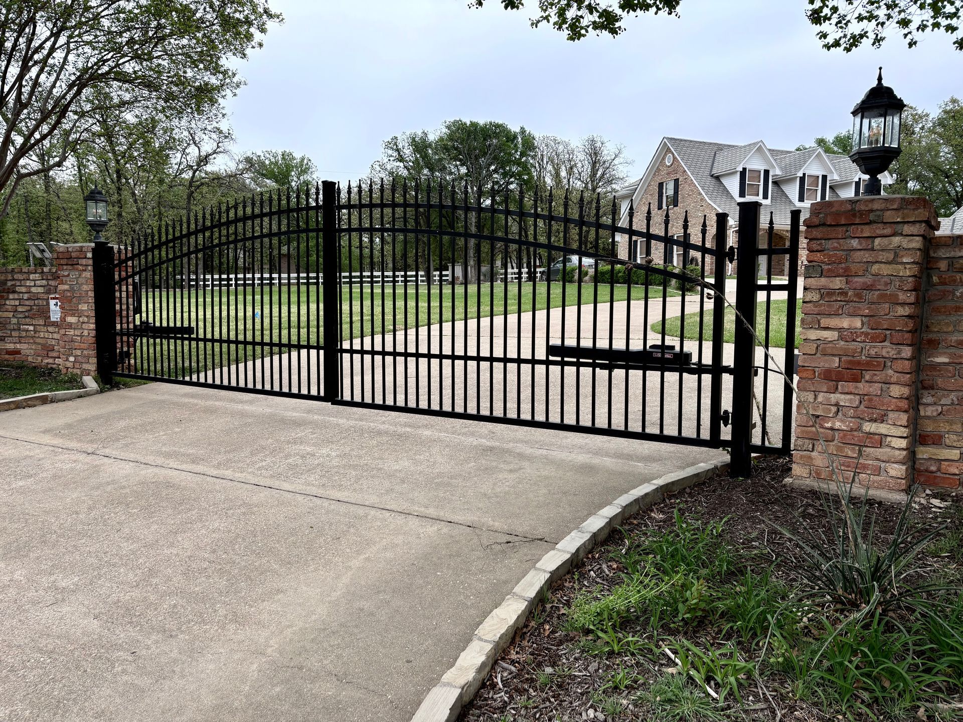 Black wrought-iron gate across a curved driveway beside brick pillars and a house behind it
