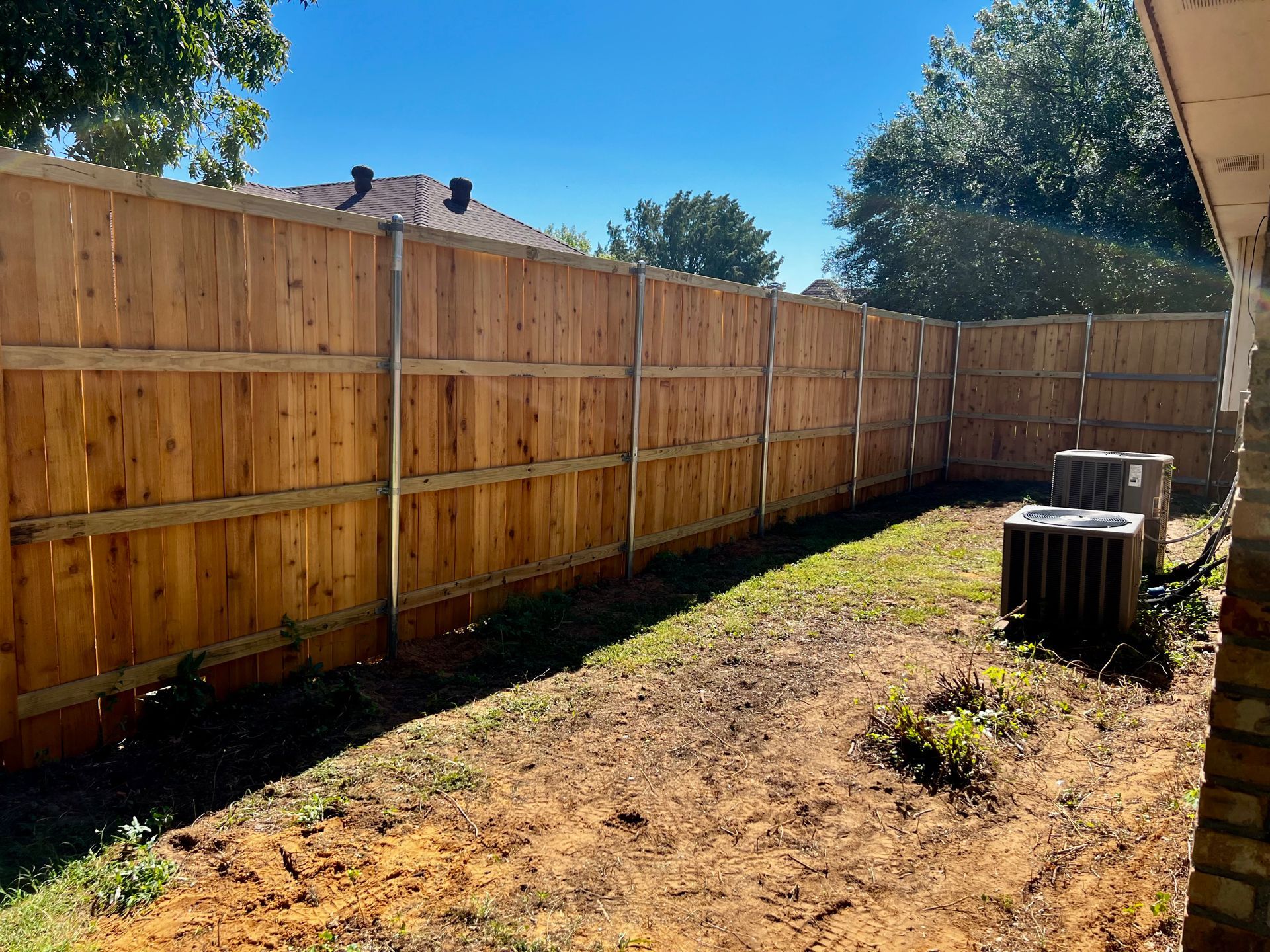 Backyard with a long wooden privacy fence, dirt path, and an outdoor AC unit in sunlight