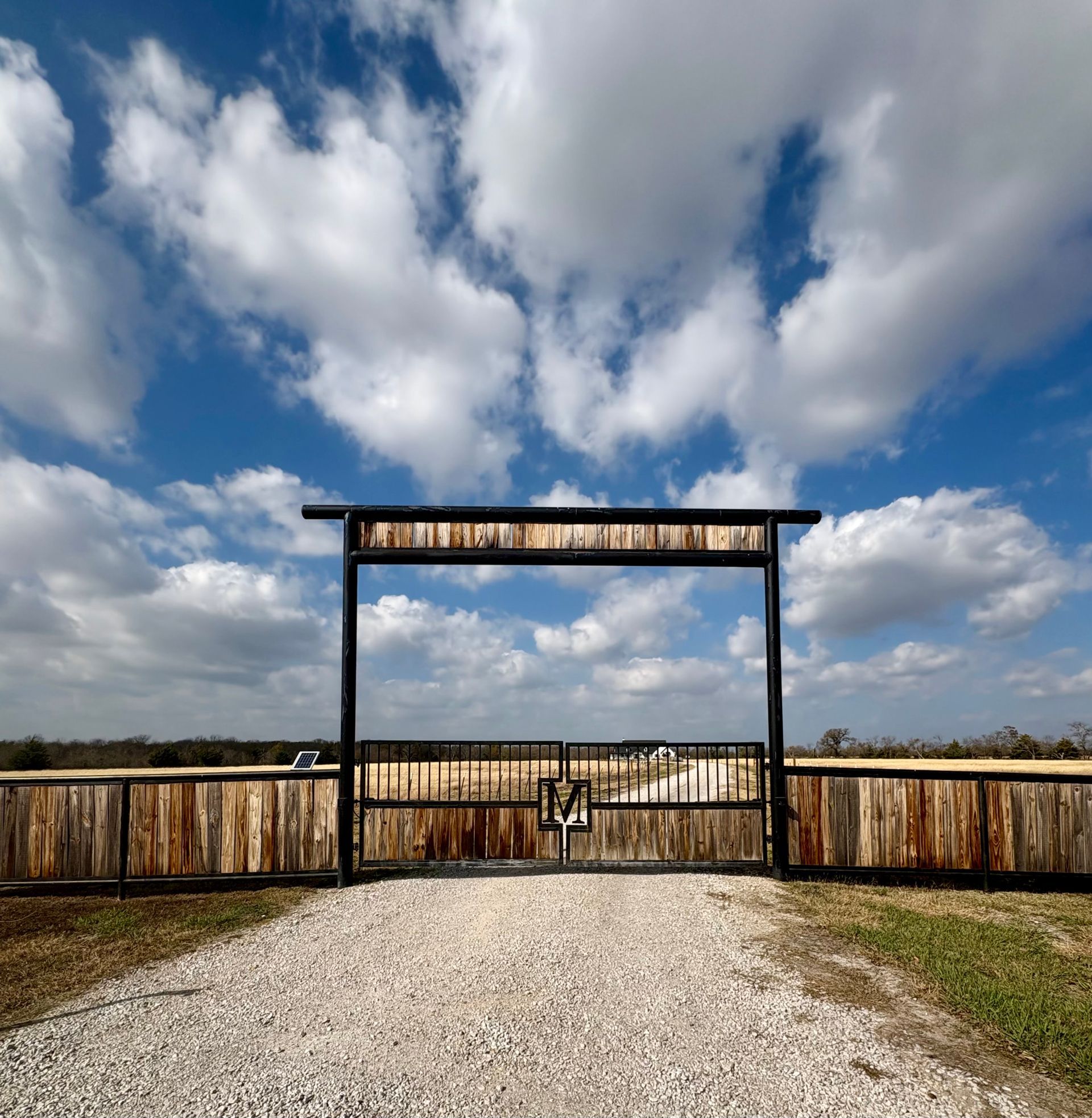 Gravel path to a wooden ranch gate under a blue sky with clouds