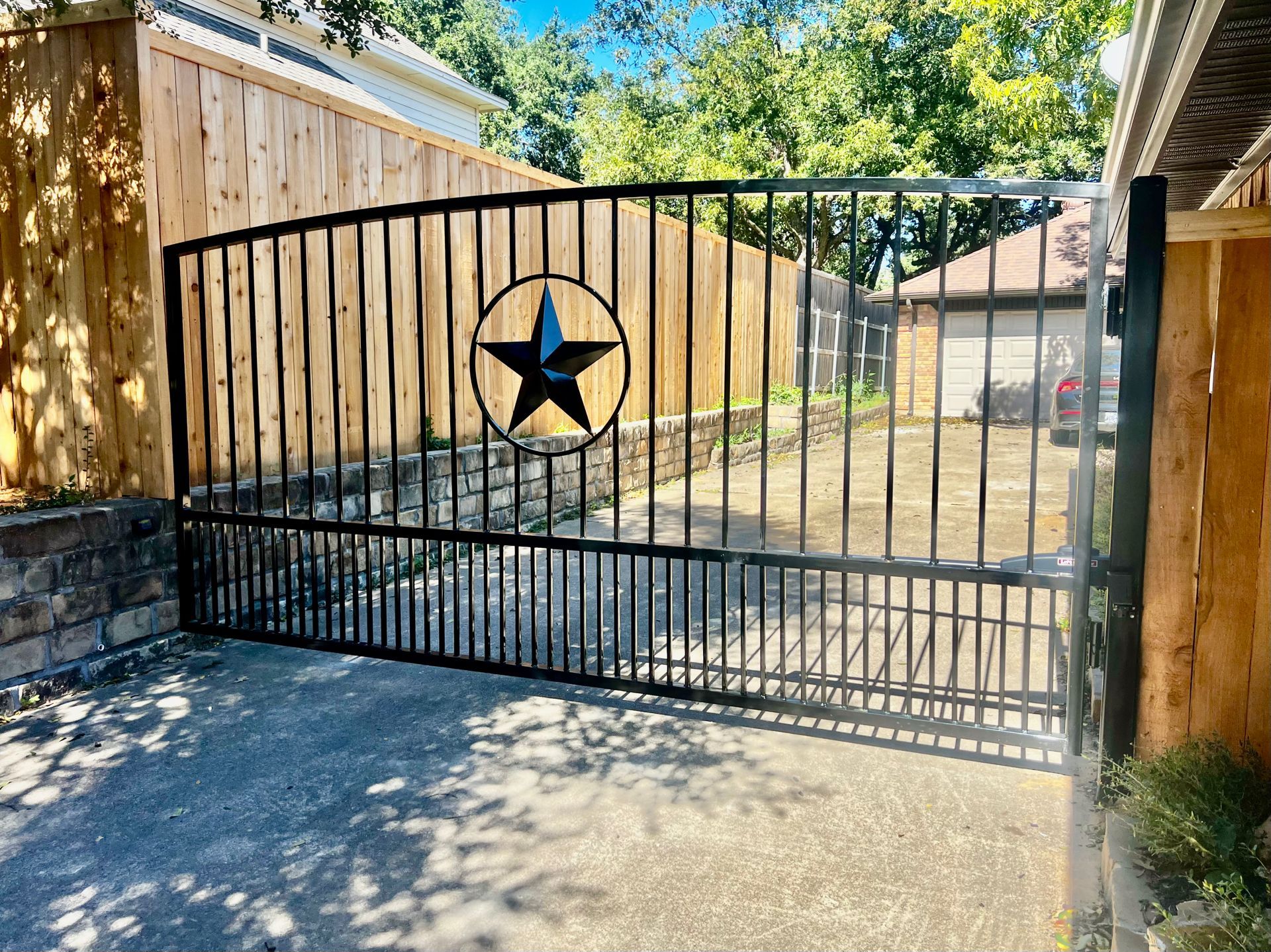 Black metal gate with a star emblem across a driveway between wooden fences.