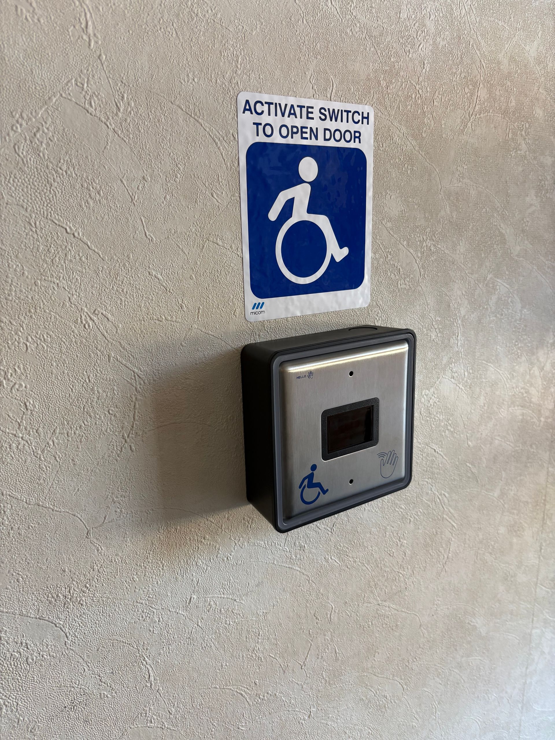 Wall-mounted accessible door switch with blue wheelchair sign and “activate switch to open door” label.