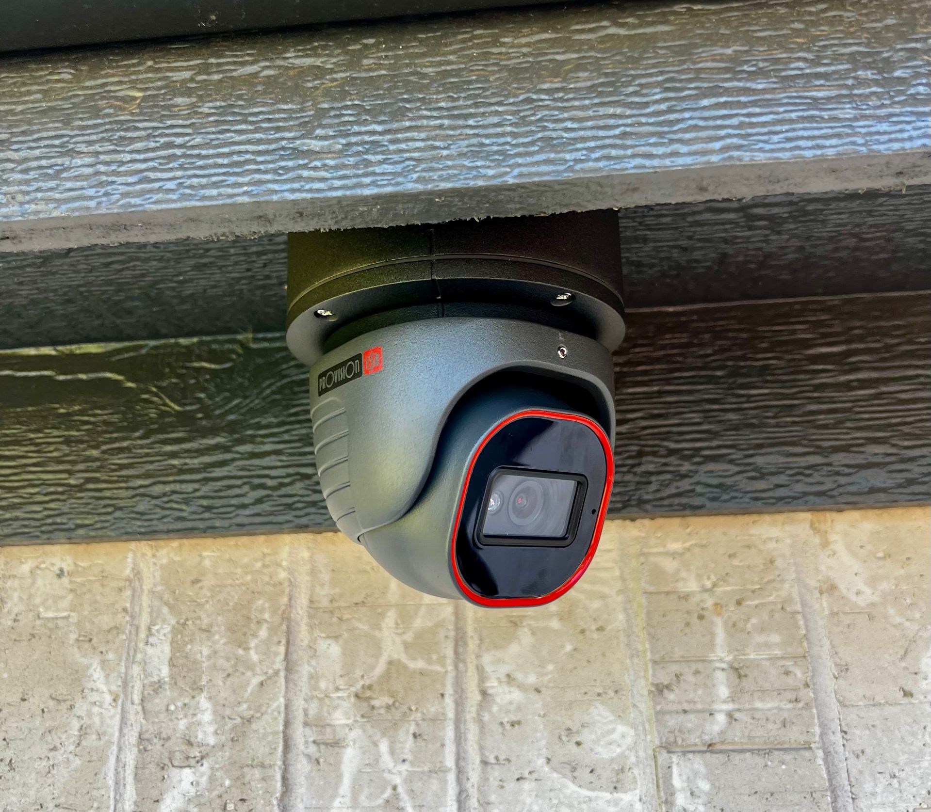 Outdoor security camera mounted under a wooden overhang, aimed downward over a stone wall