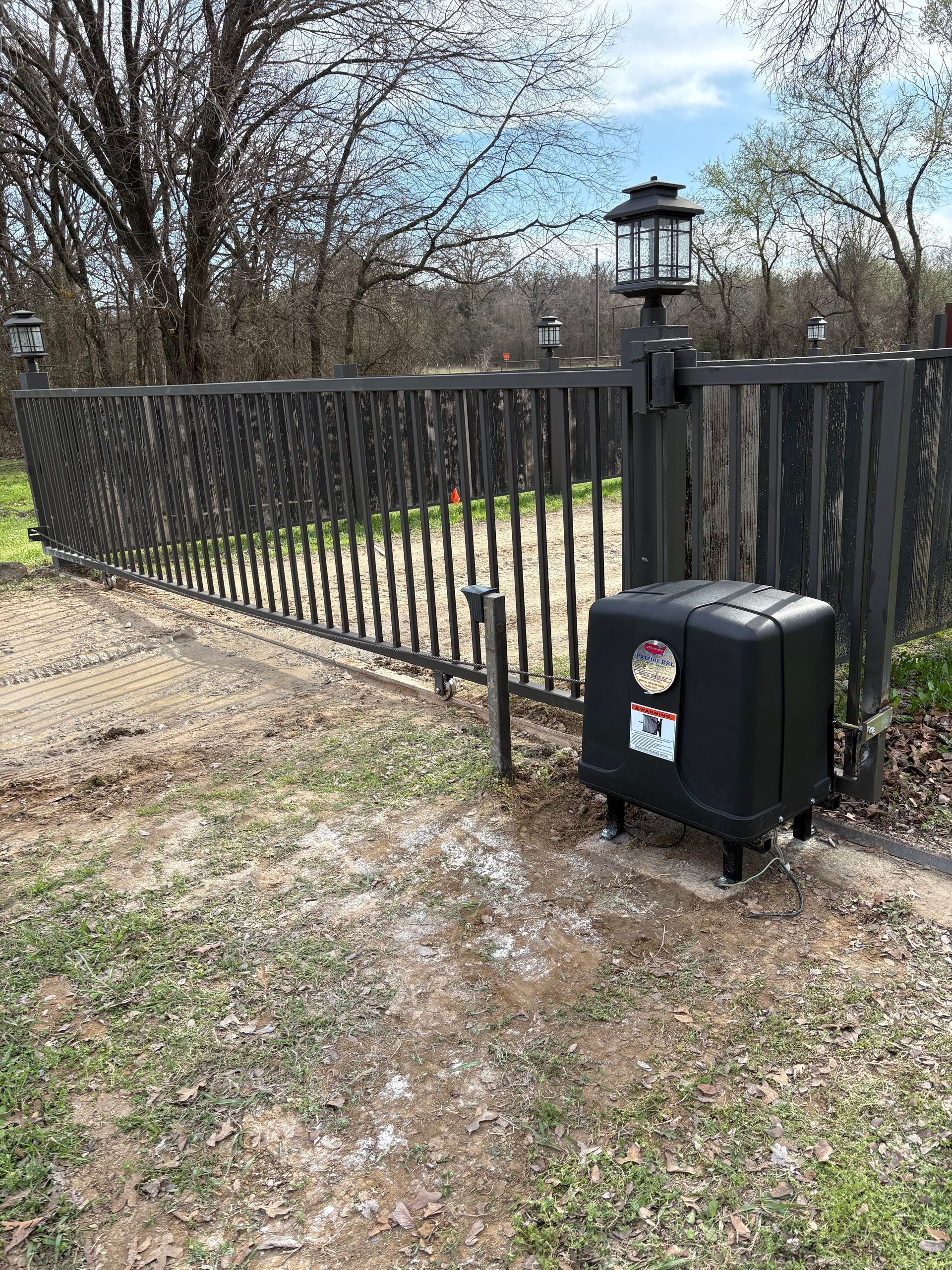 Black metal gate and fence beside a lantern post, with a small utility box on a gravel path in a yard.