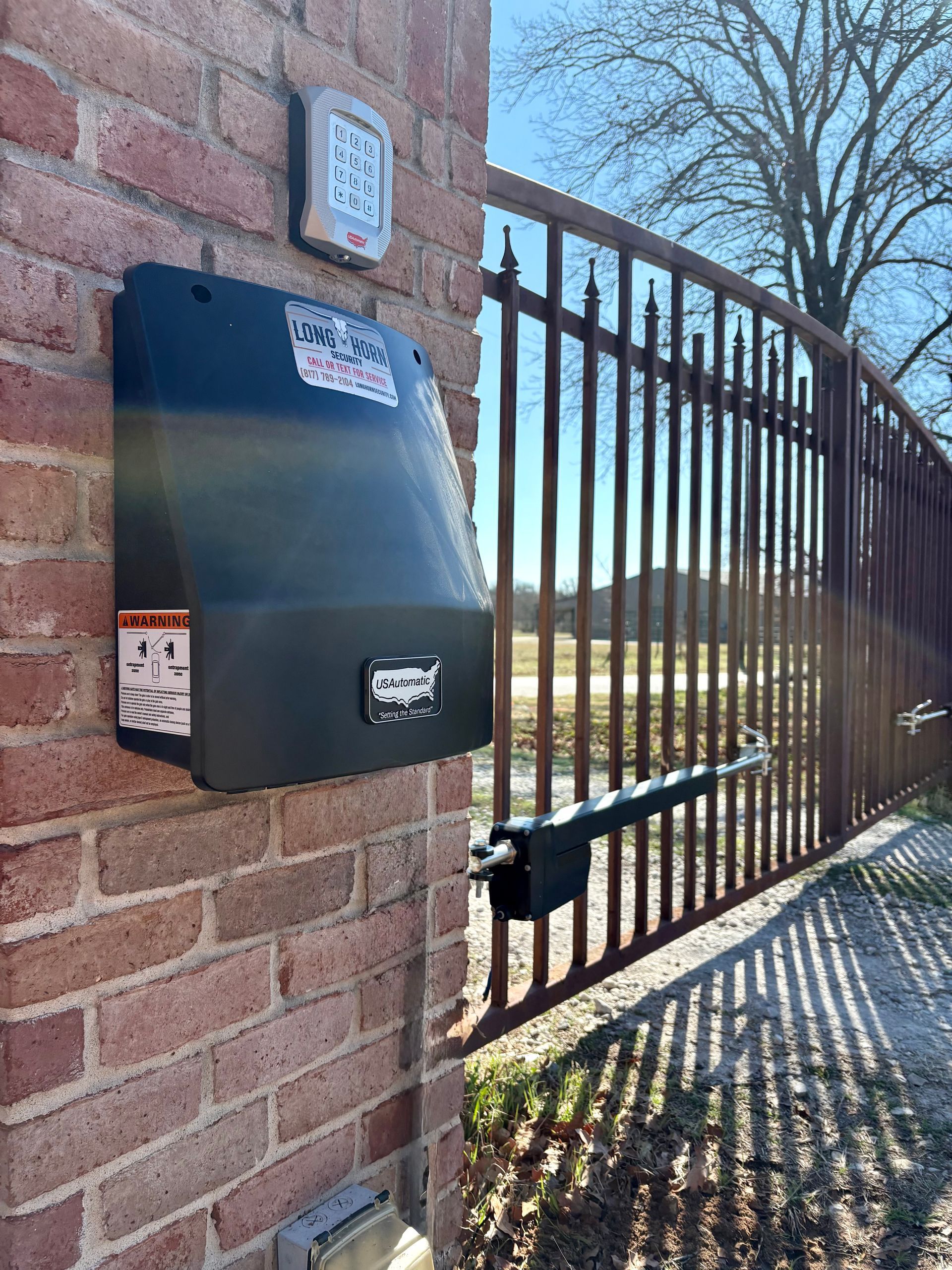 Black wall-mounted mailbox on a brick wall beside a metal gate in sunlight
