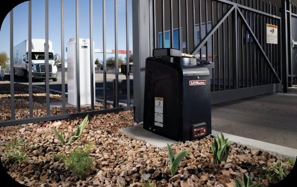 Black street-side utility unit by a metal gate, beside gravel and small green plants.