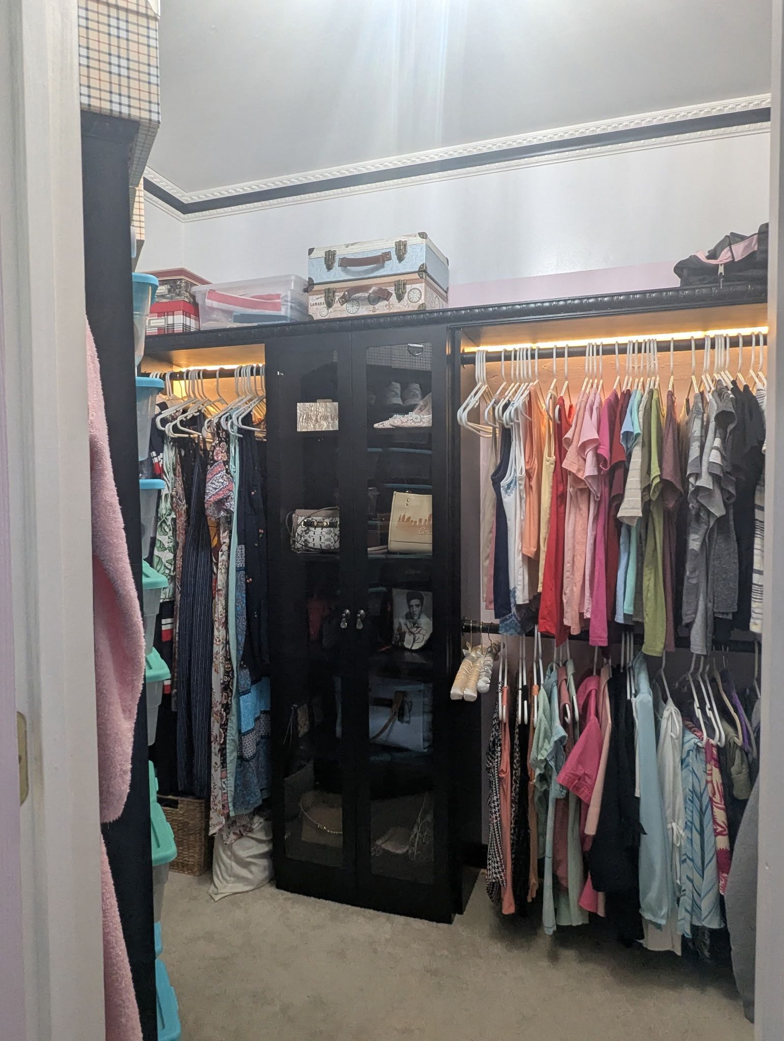 Closet with hanging clothes, shelves, and a black storage cabinet in a walk-in room