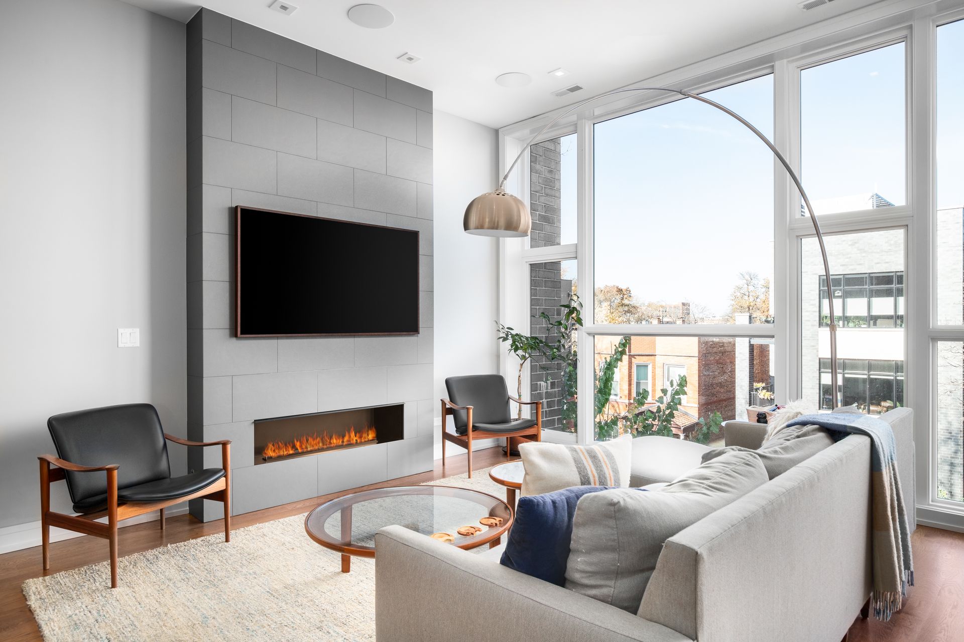 Modern living room with gray sofa, wall-mounted TV, fireplace, and large windows overlooking a city view