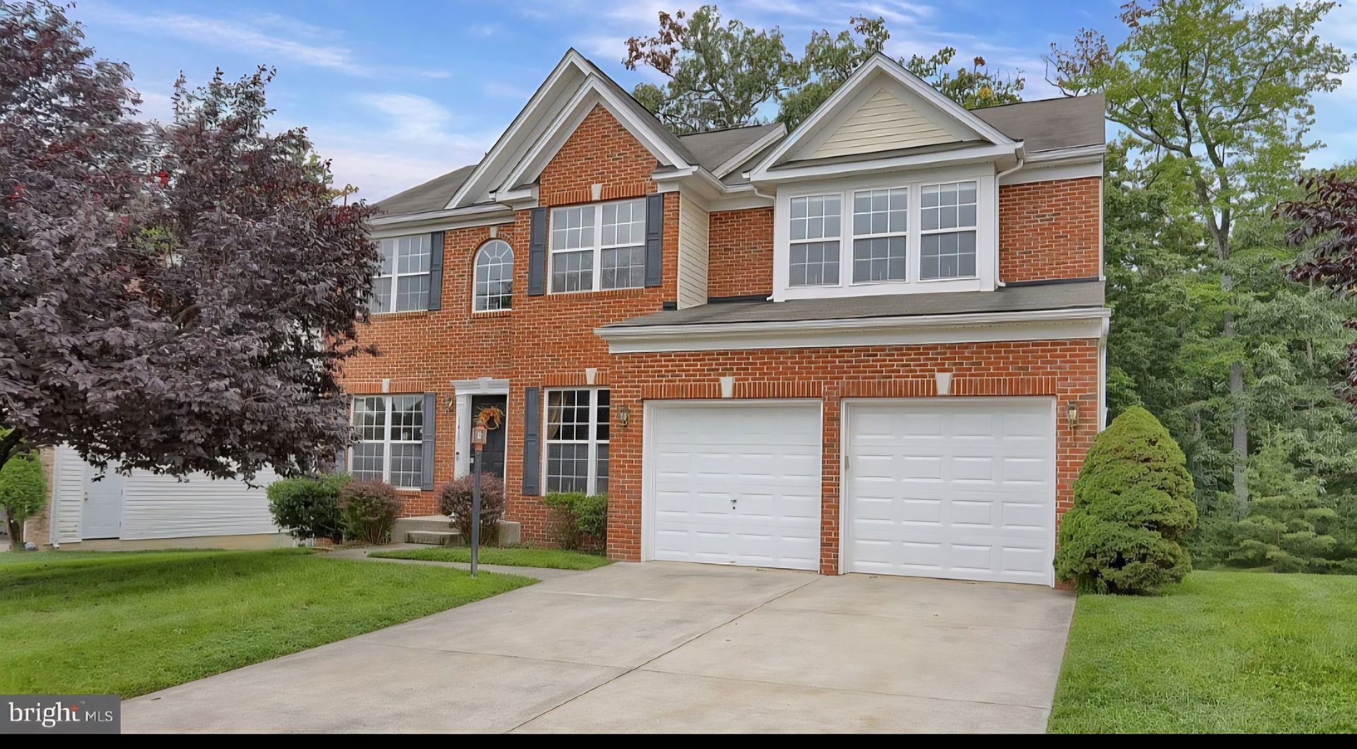 Two-story brick suburban house with white garage doors, driveway, and trees in the front yard