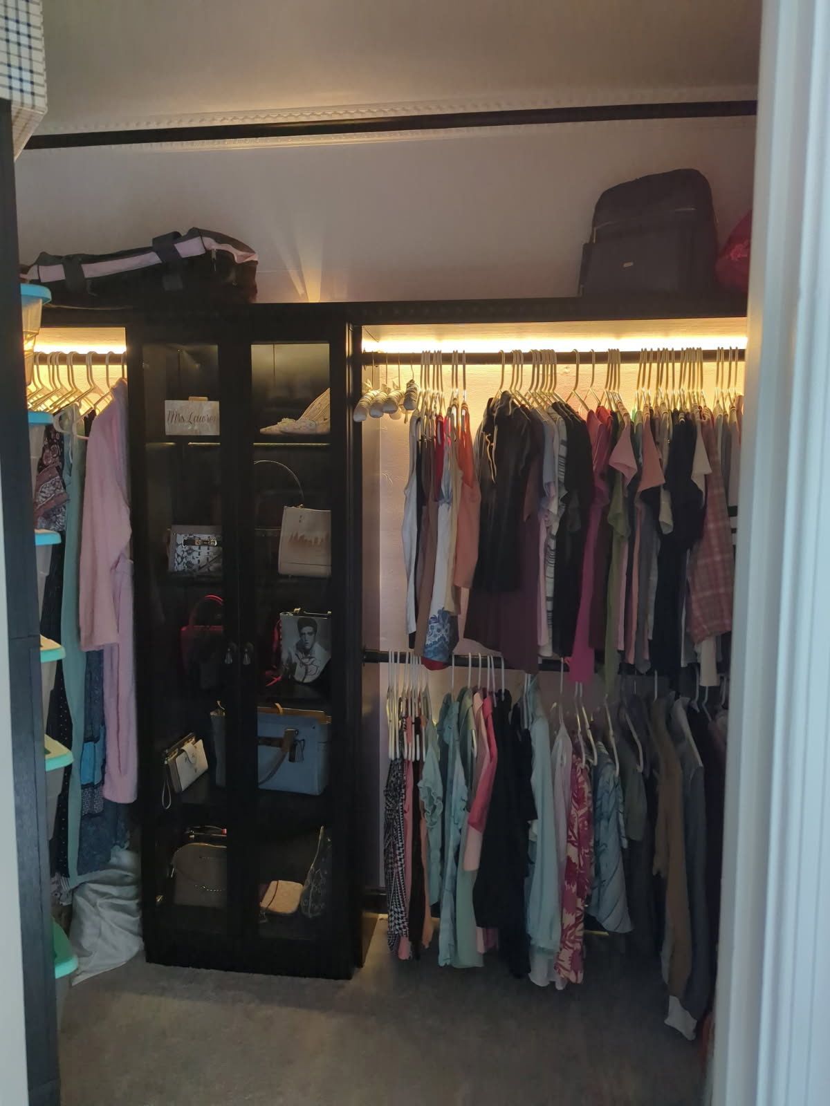 Lit closet with hanging clothes, shelving, and a suitcase on the top shelf