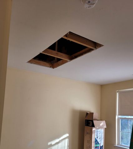 Room ceiling with a large rectangular hole exposing wooden framing near a window.