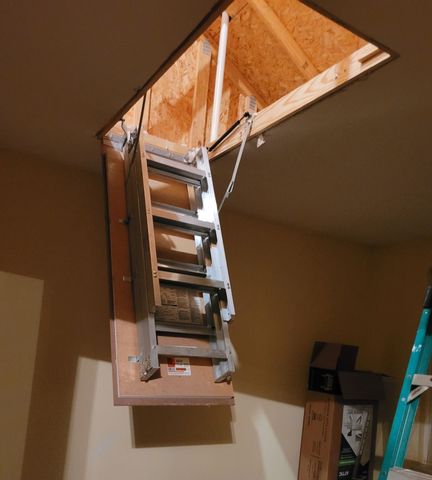 Open attic hatch with ladder extended into a beige room, revealing plywood insulation above.