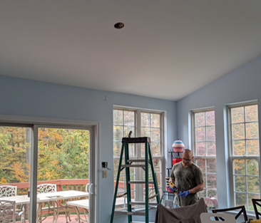 Two people working in a bright room with a ladder and windows, near a deck outside.