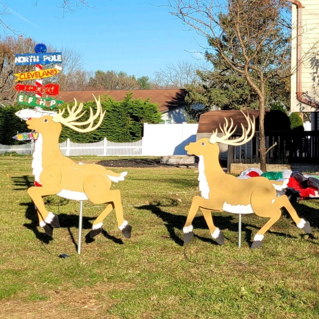 Two cardboard deer lawn decorations in a sunny yard with a house and trees behind them.
