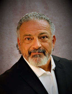 Portrait of a man in a black suit and white shirt with gray hair and beard, against a dark studio backdrop