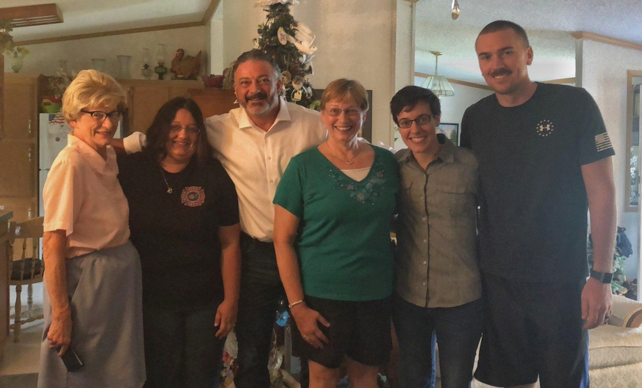 Six people standing together indoors in front of a Christmas tree, smiling at the camera