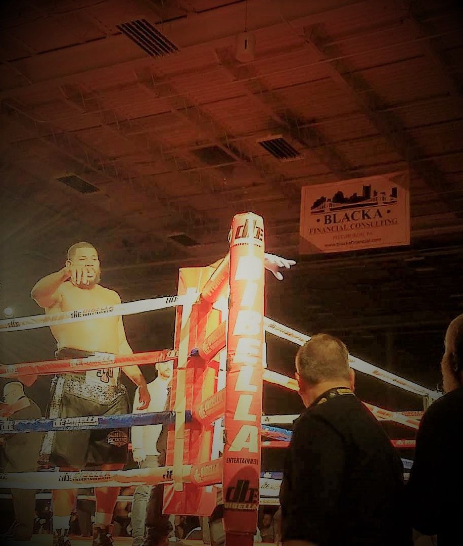 Boxing ring with two fighters under warm lights, one in red shorts, spectators visible around the ropes