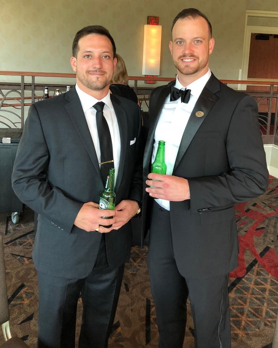 Two men in black tuxedos holding green bottles at an indoor event.