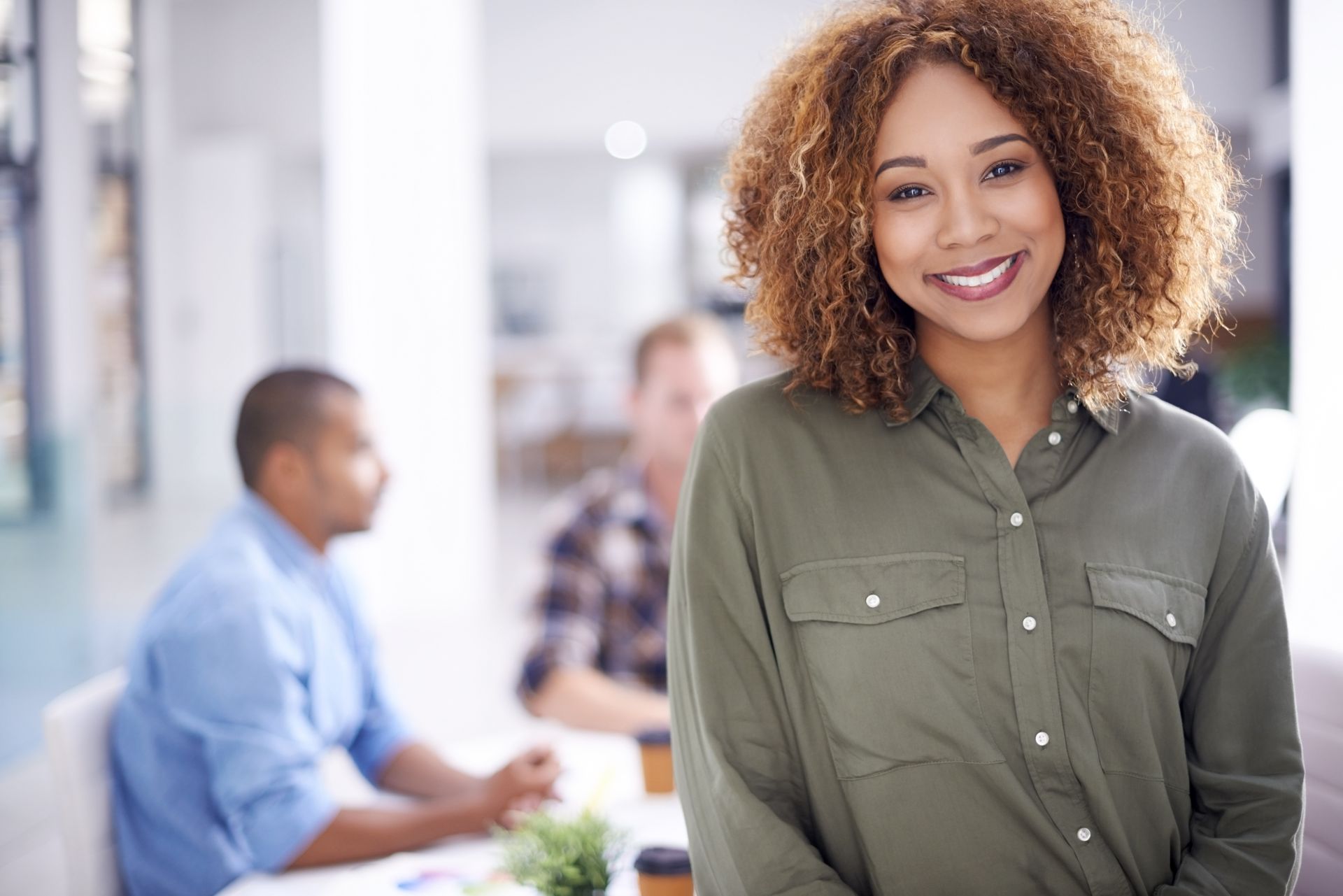Smiling woman in an olive green shirt stands in an office with coworkers blurred in the background