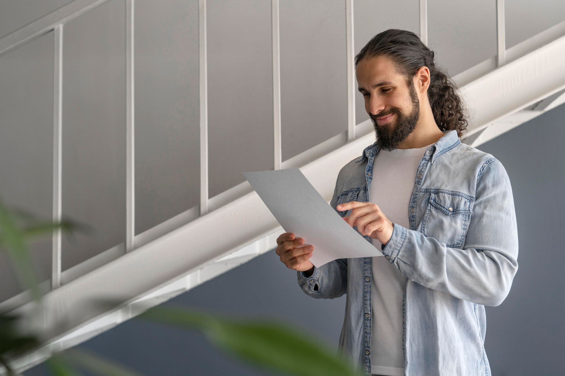 Person smiling and reading a document in a bright modern hallway, wearing a denim jacket.