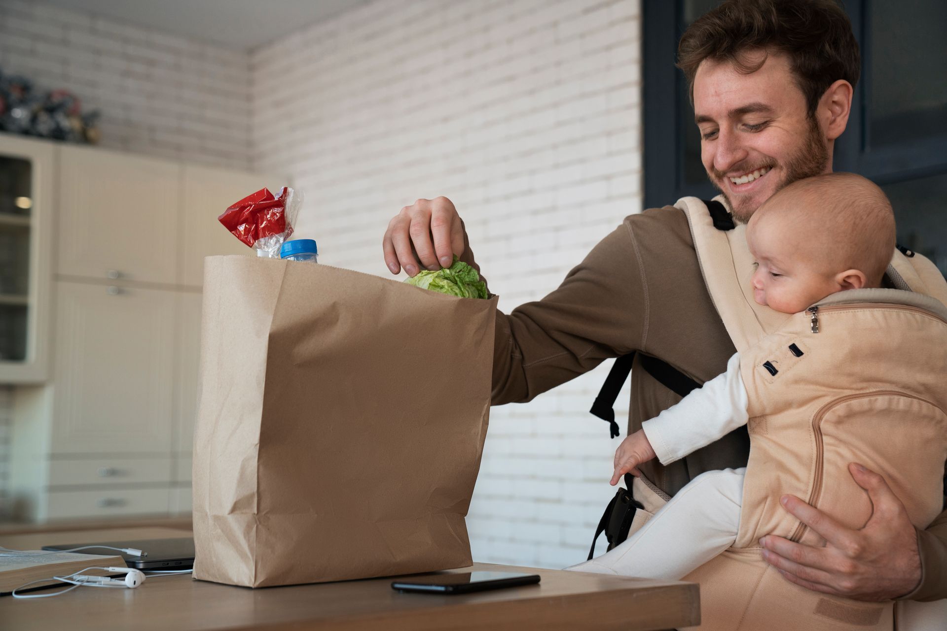 Man smiling while holding a baby beside a grocery bag on a kitchen table