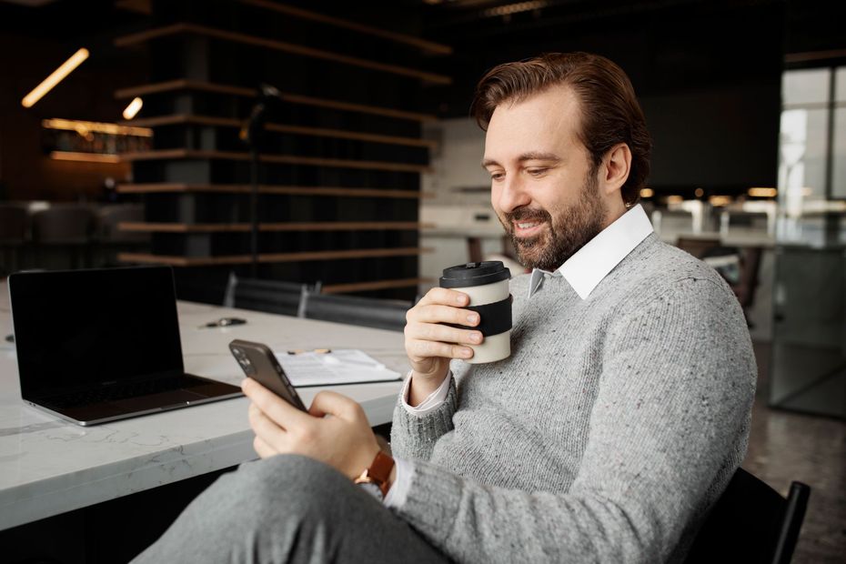 Man sitting in a café, smiling at his phone while holding a takeaway coffee cup.