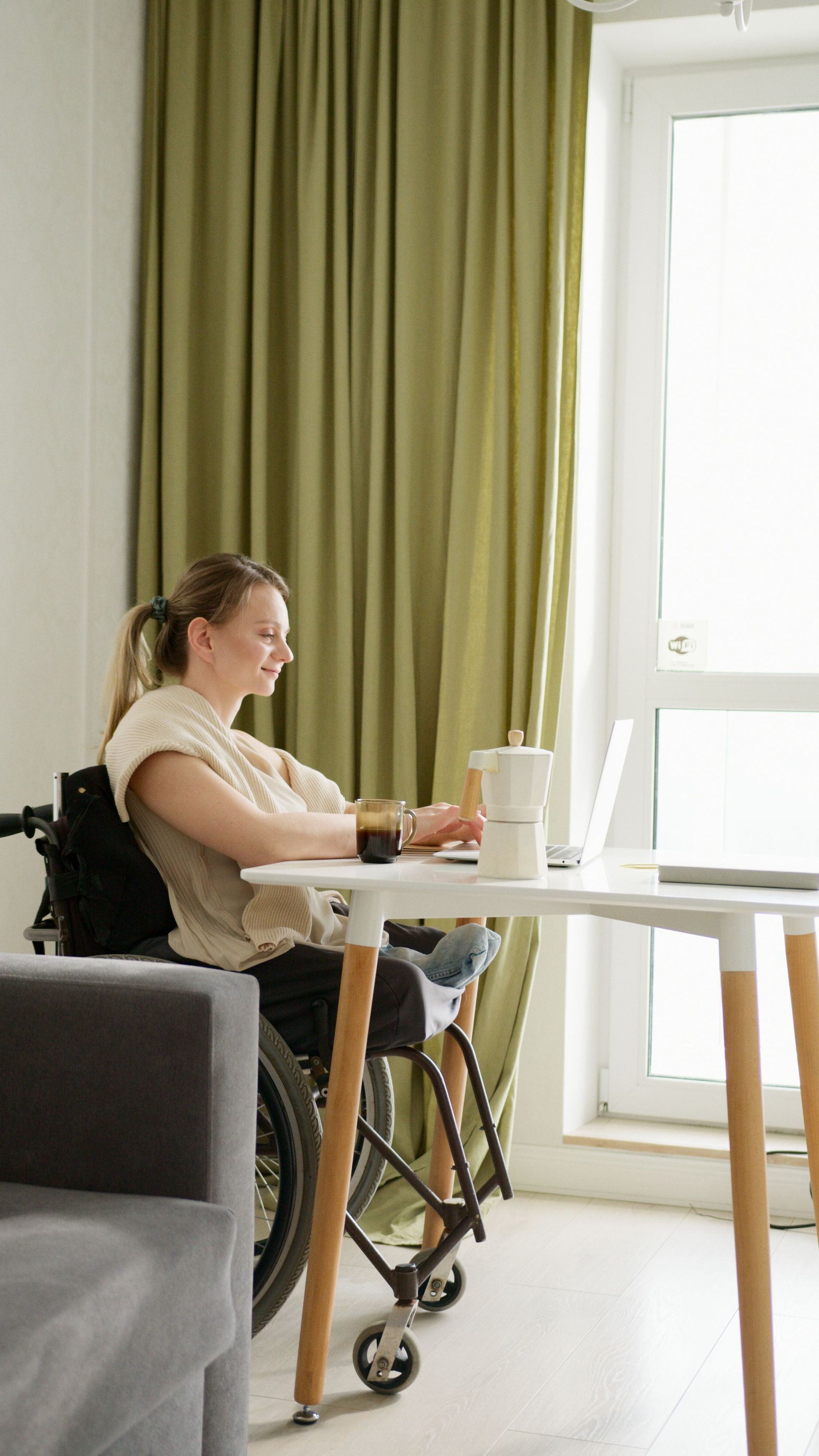 Person seated at a table by a bright window, looking at a laptop in a cozy room with green curtains.