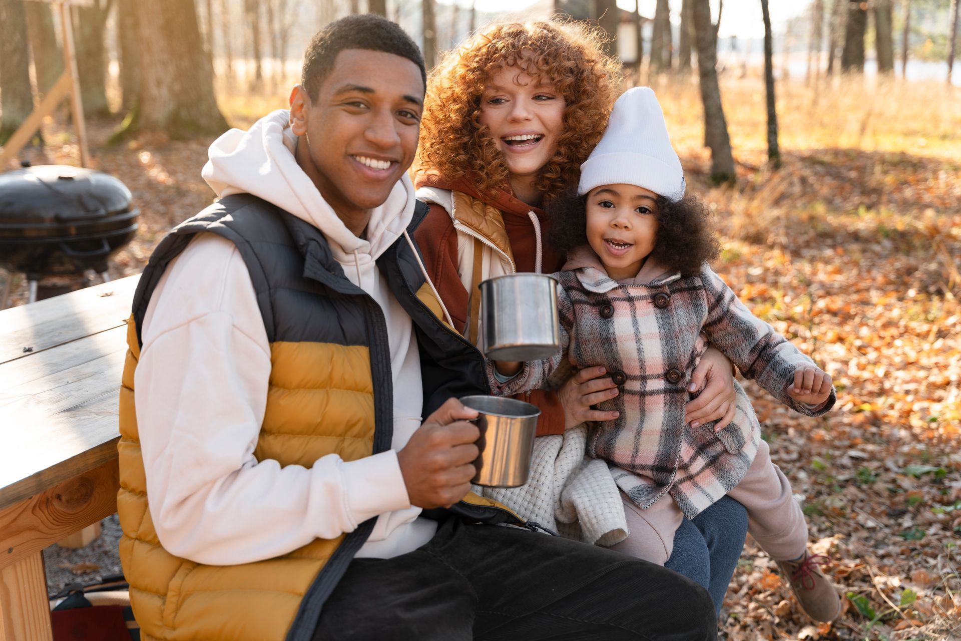Family sitting on a park bench, smiling and holding mugs in a sunny autumn setting.