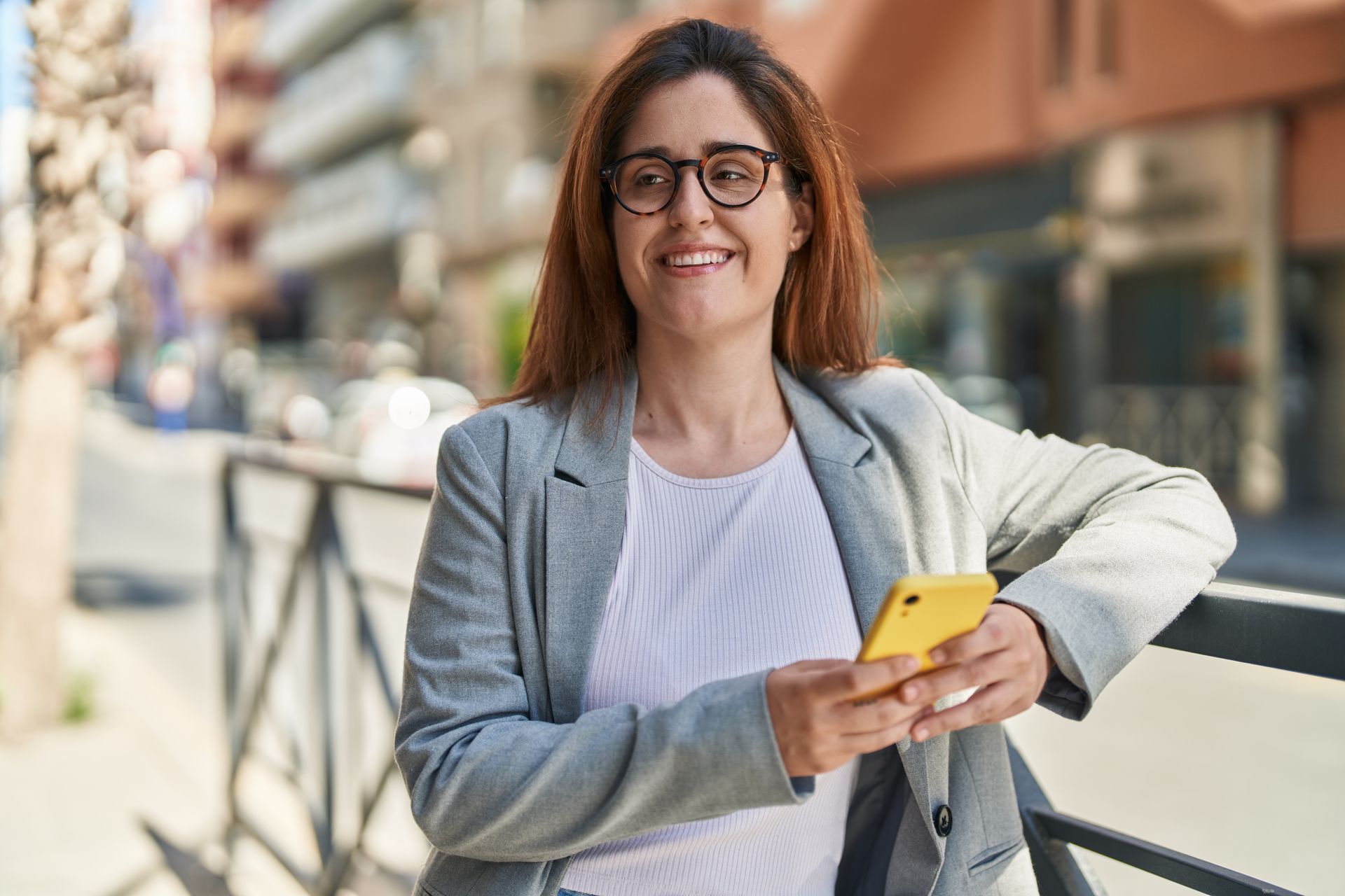 Woman smiling on a city street, holding a yellow phone and wearing a gray blazer.
