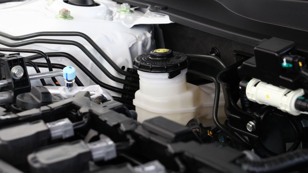 Car engine bay with fluid reservoirs, black hoses, and wiring near the firewall.