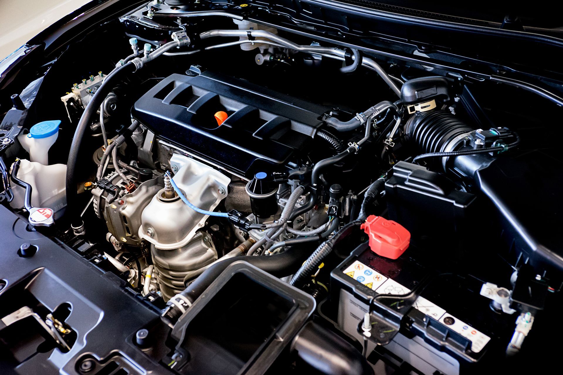 Car engine bay with black cover, silver components, and a red battery terminal