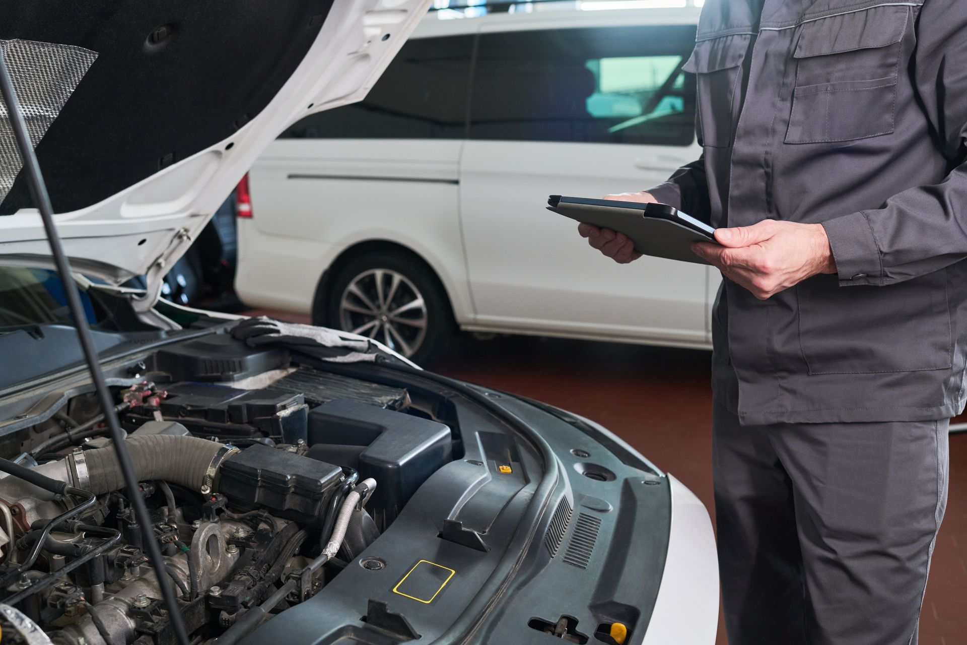 Mechanic inspecting a car engine with hood open, holding a tablet in a garage.