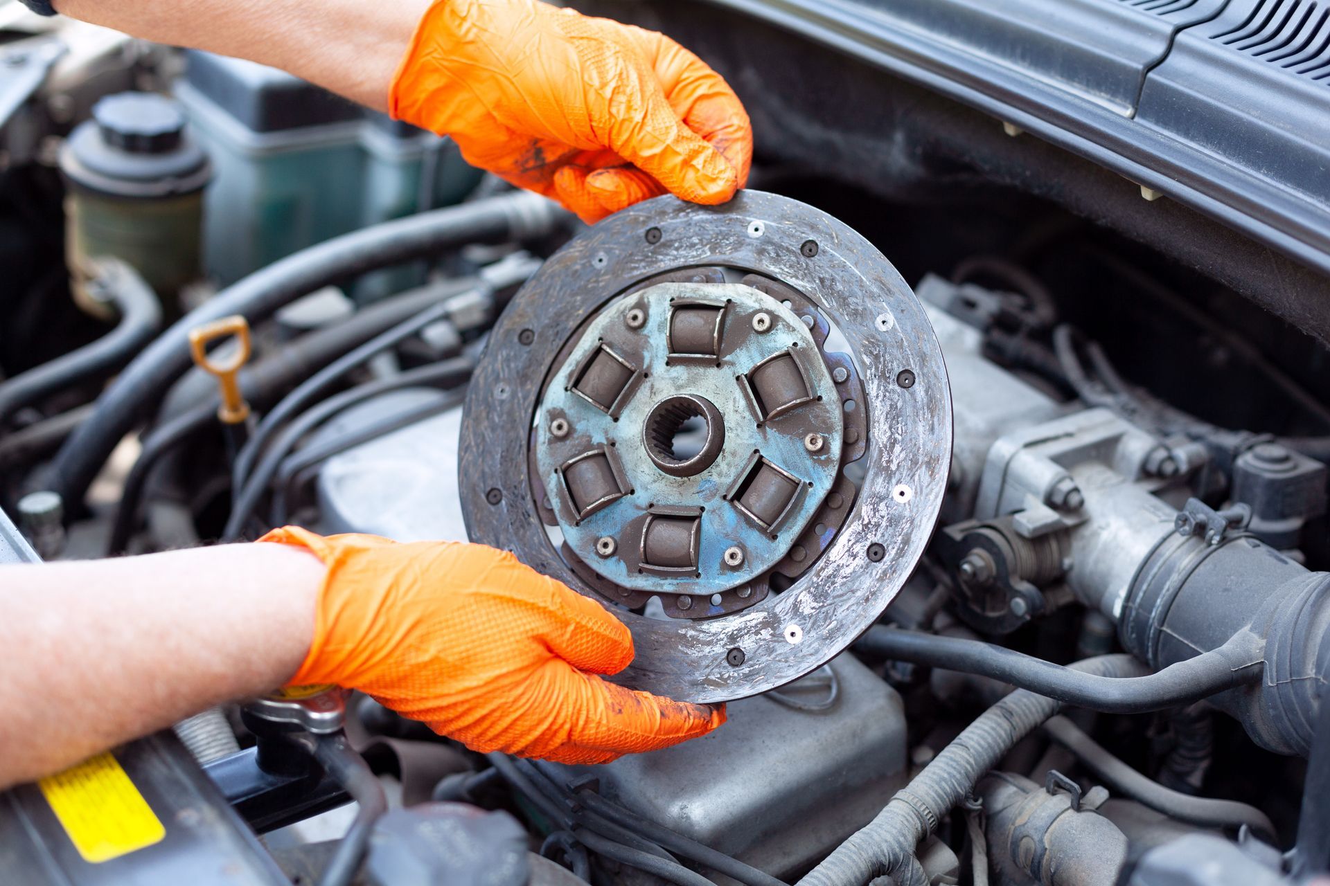Gloved hands holding a worn car clutch disc inside an open engine bay Gloved hands holding a worn car clutch disc inside an open engine bay