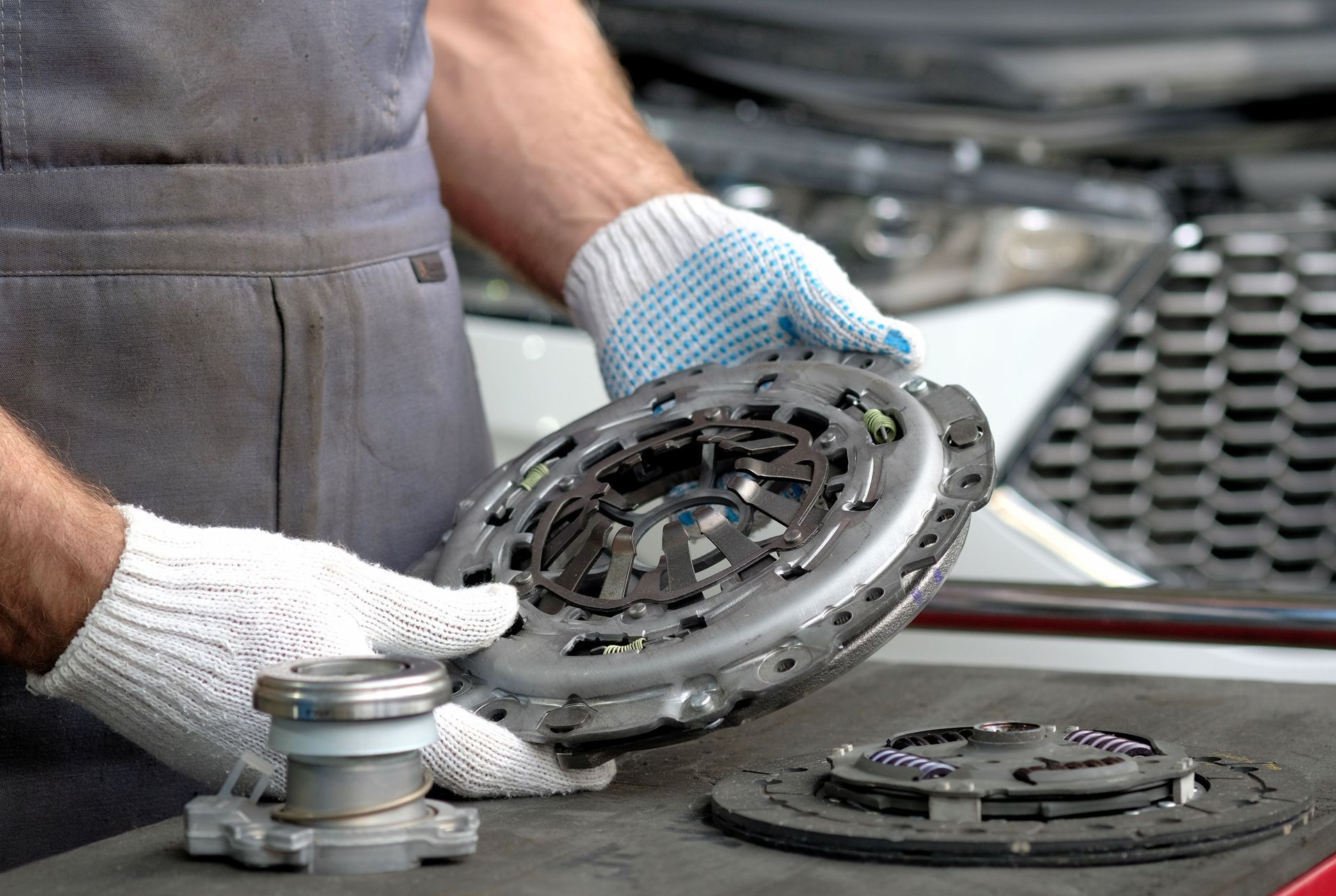 Mechanic wearing gloves holding a car clutch assembly over a workbench with parts in the background Mechanic wearing gloves holding a car clutch assembly over a workbench with parts in the background