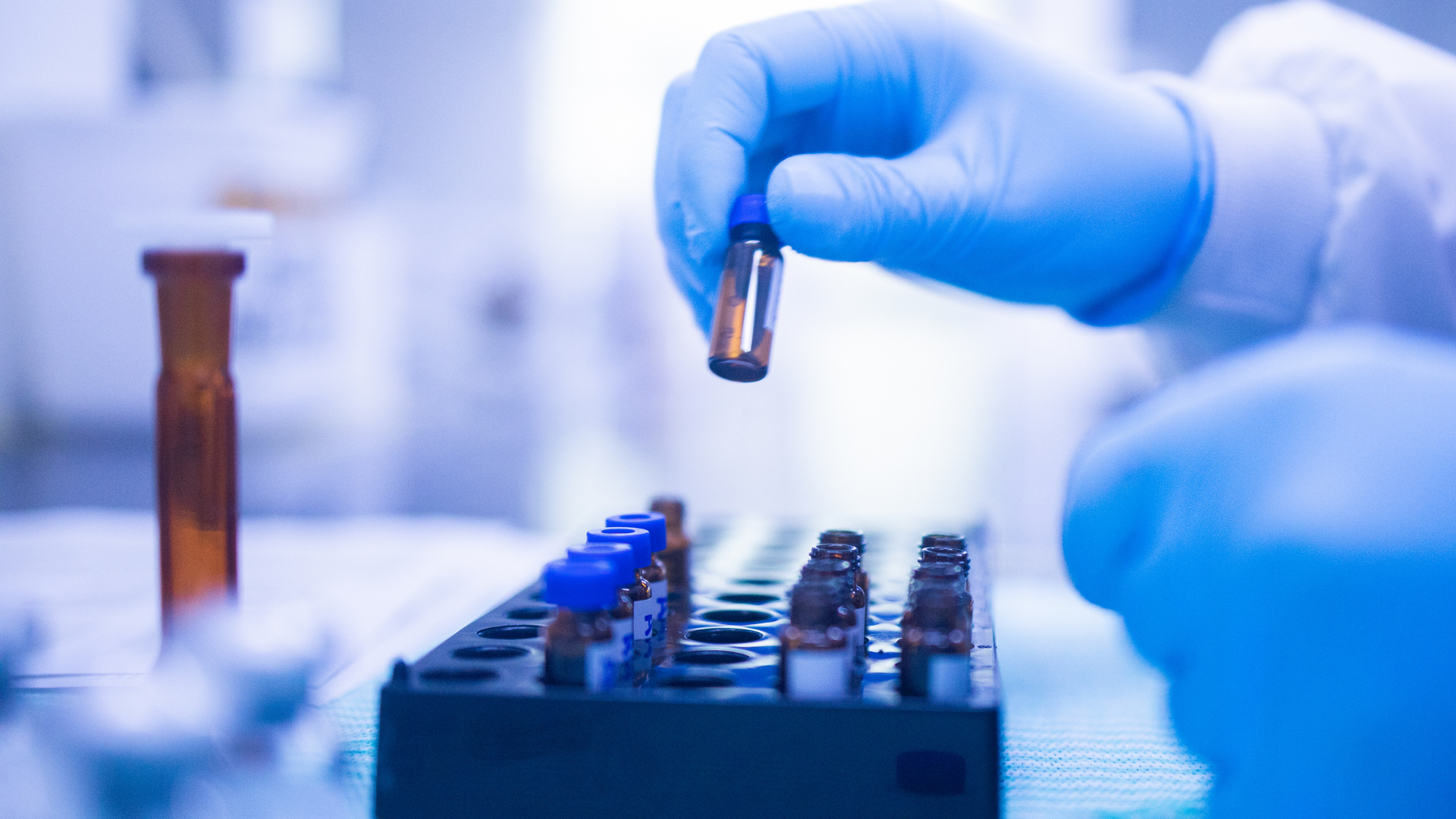 Blue-gloved hand holding a test tube above a lab sample rack in a clinical setting