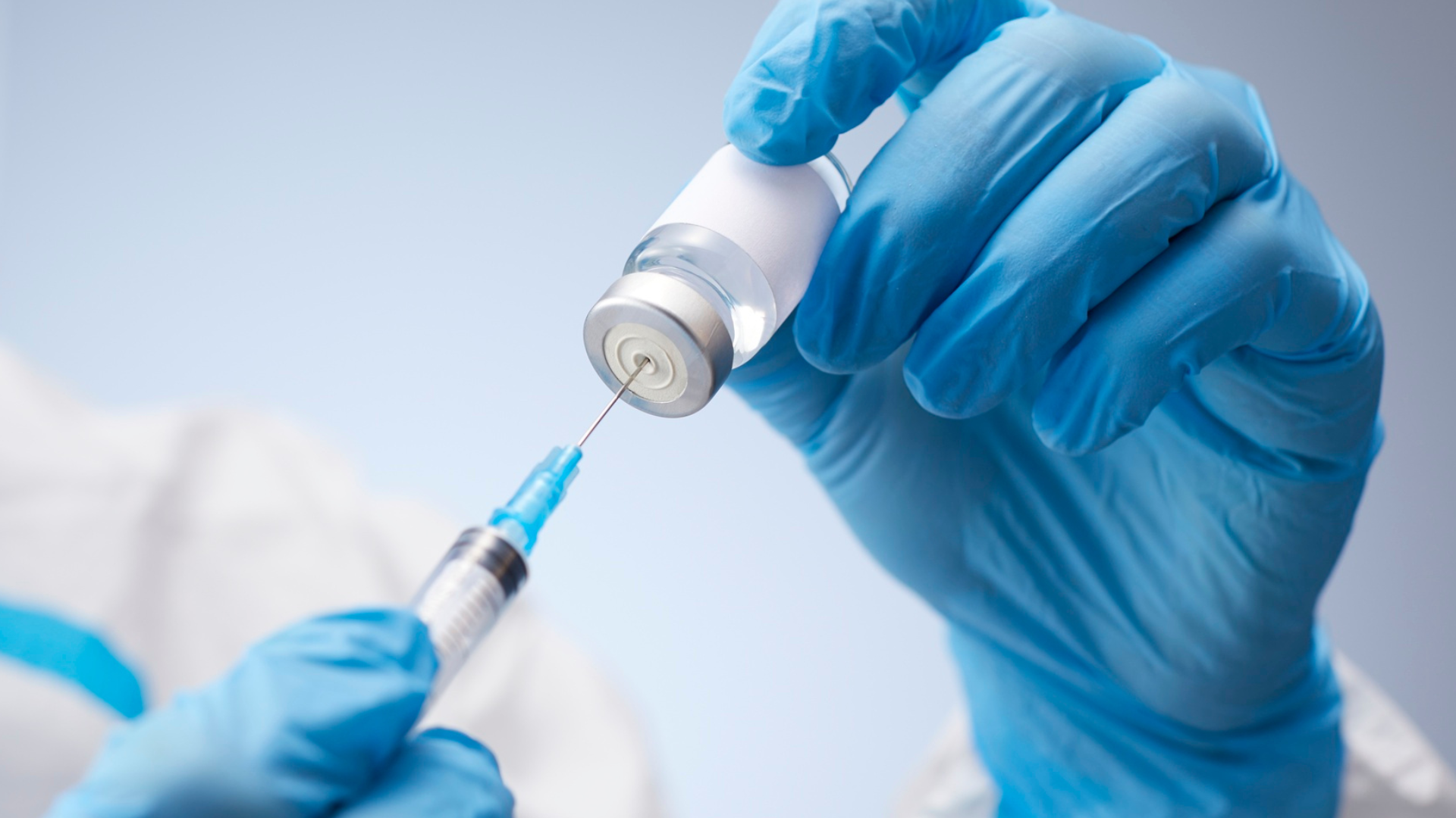 Gloved hands drawing vaccine from a vial into a syringe in a clinical setting