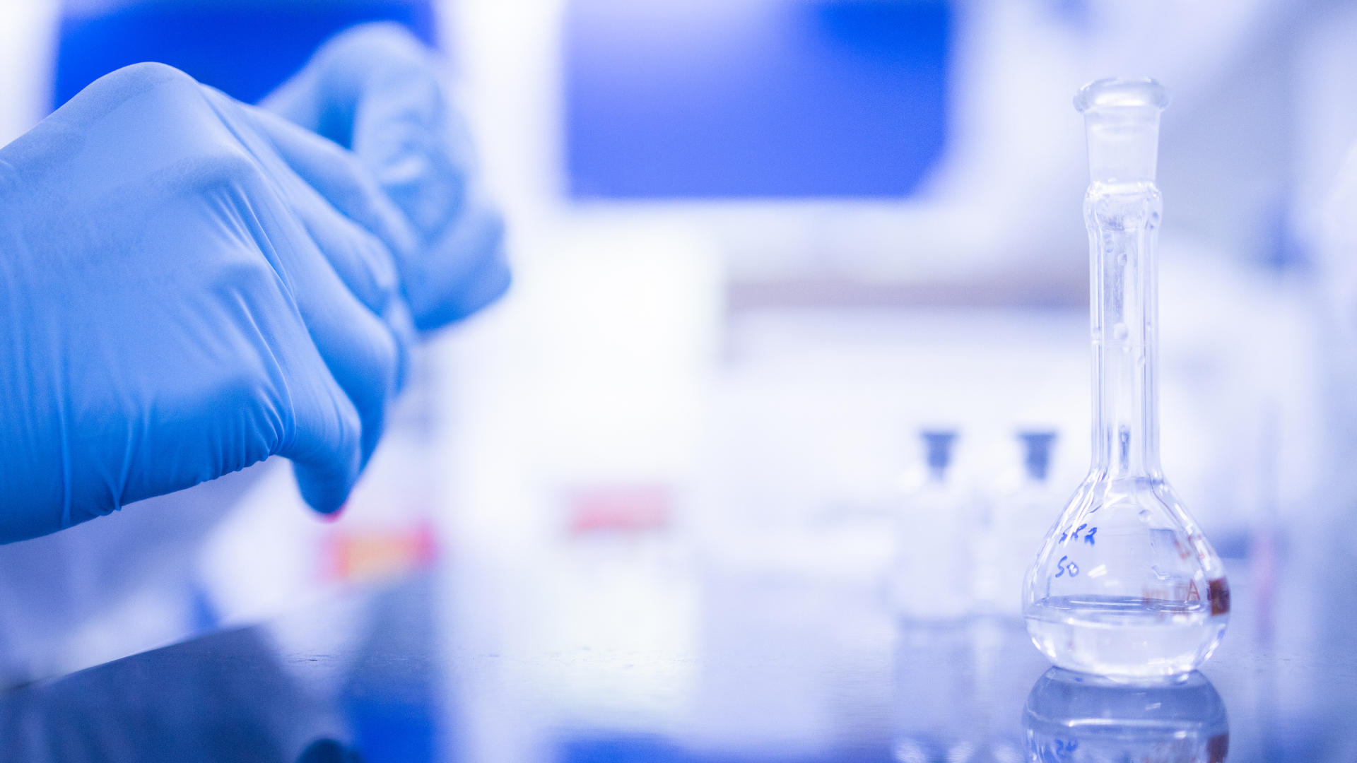 Gloved hand holding a pipette in a blue-lit laboratory with glass labware on the bench