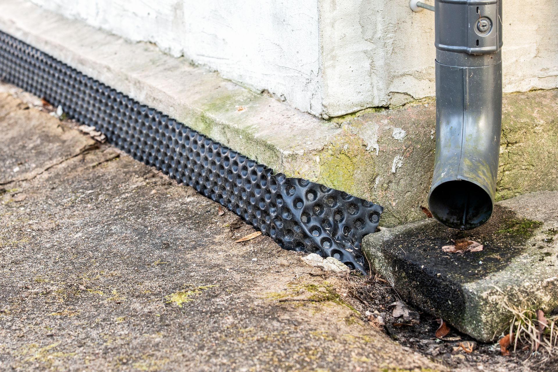 Black gravel guard and downspout at the base of a white wall on a concrete path