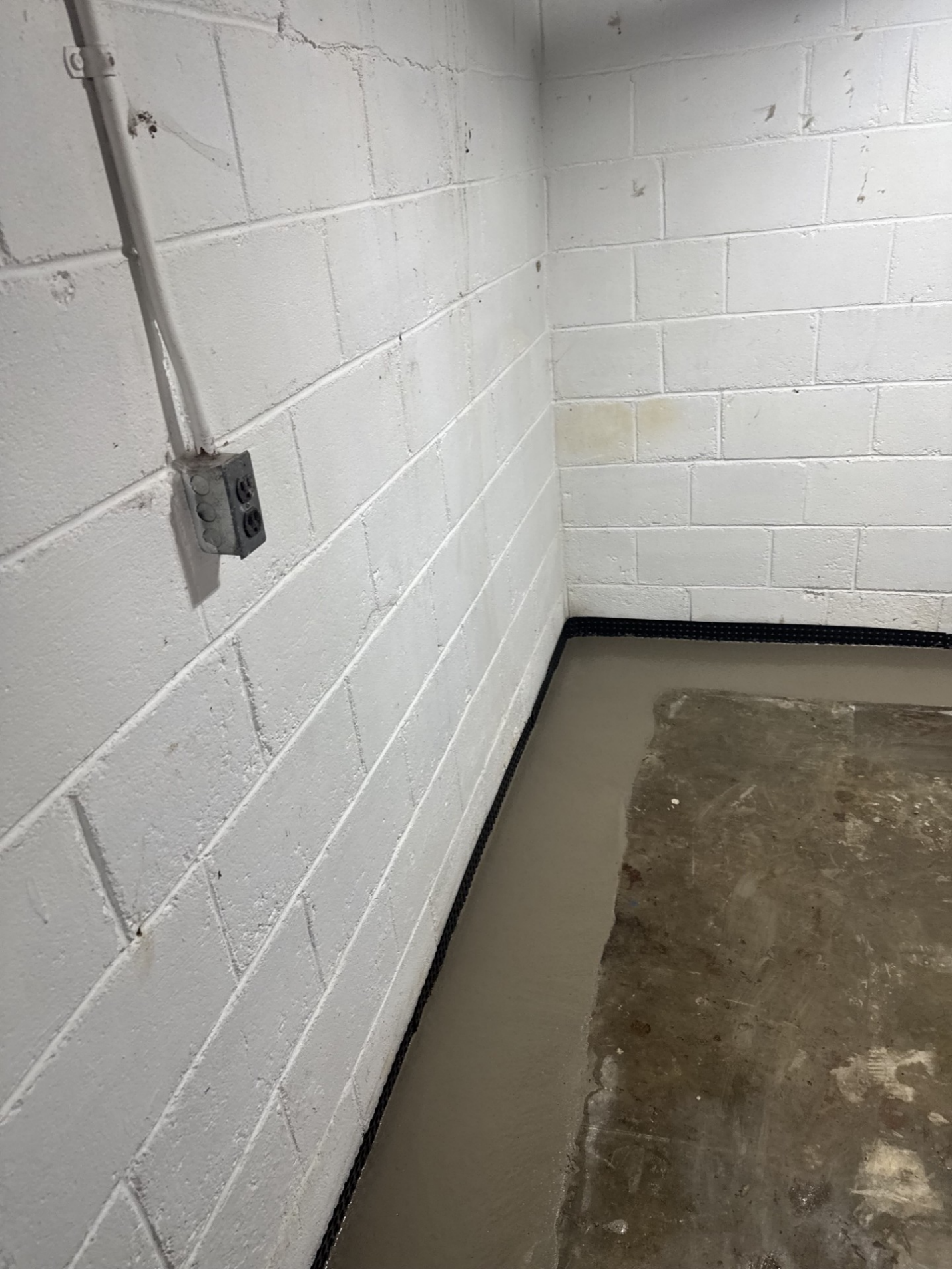 Empty cinderblock room with gray floor and a wall outlet on the left.