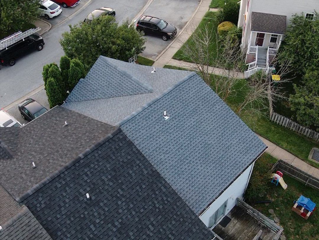 Overhead view of a house with multiple roof sections covered in dark gray shingles. Cars and a yard are in the background.