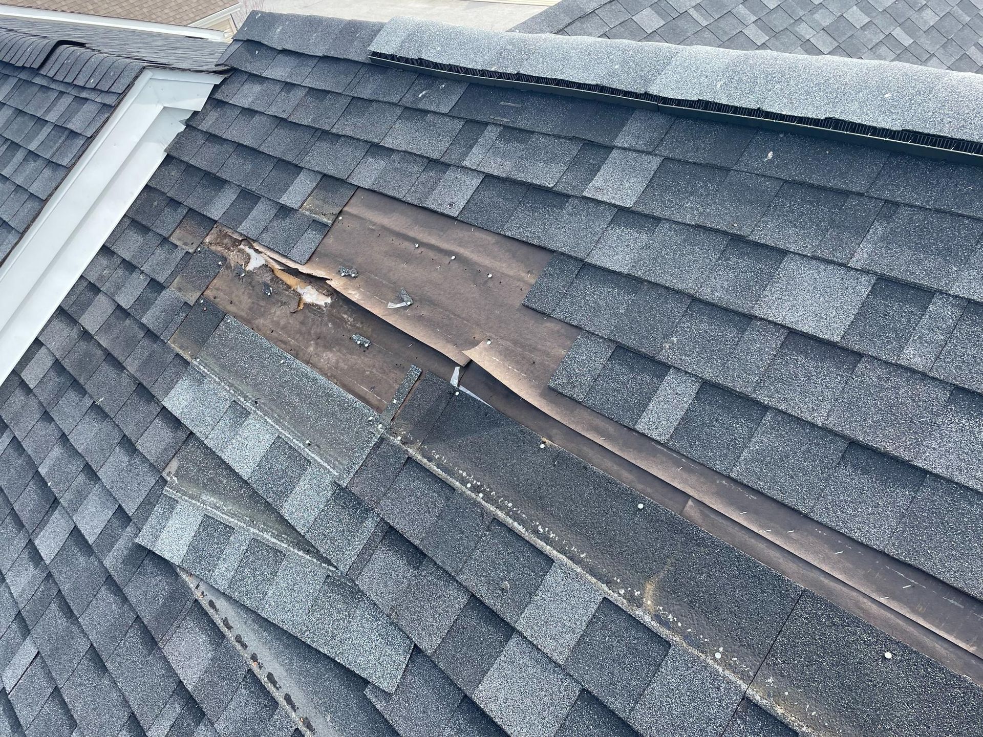Damaged asphalt shingle roof, torn shingles reveal underlayment along a roof valley, gray and tan colors.