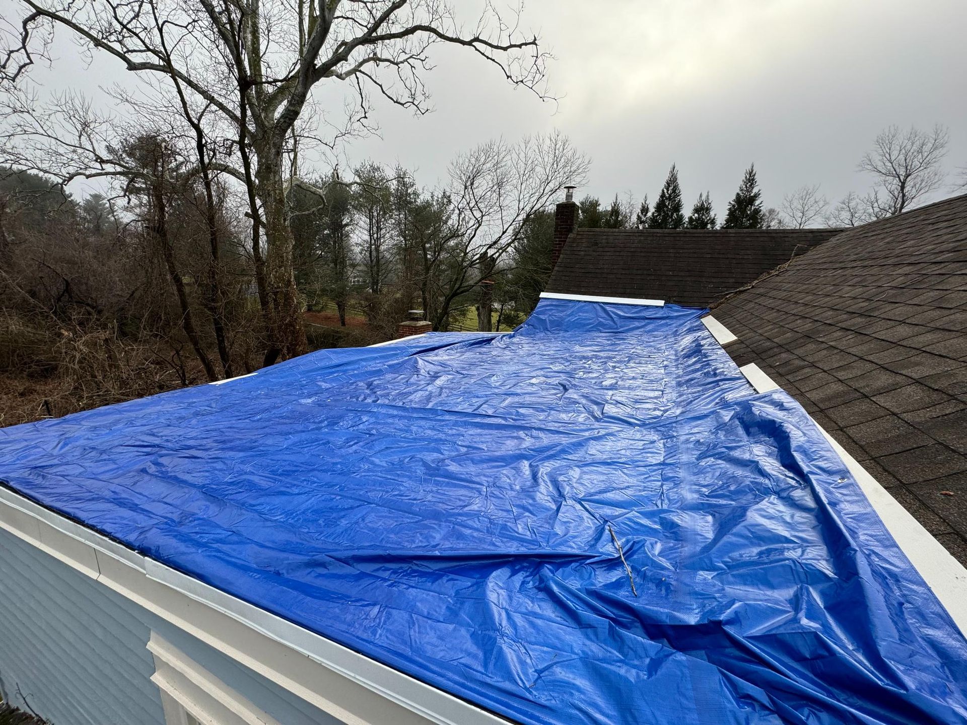 Blue tarp covers a roof, protecting a section from the elements. Overcast sky, trees in the background.