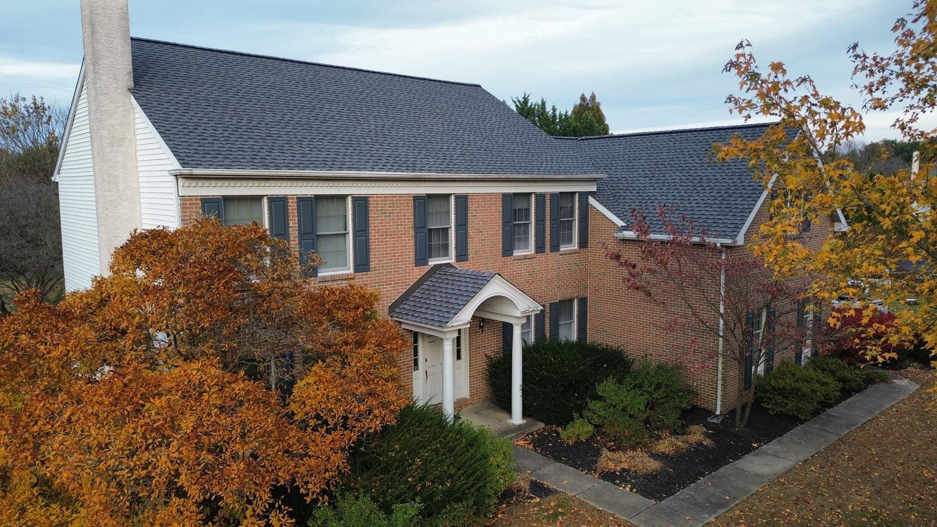 Brick house with dark roof and white chimney, surrounded by autumn foliage.