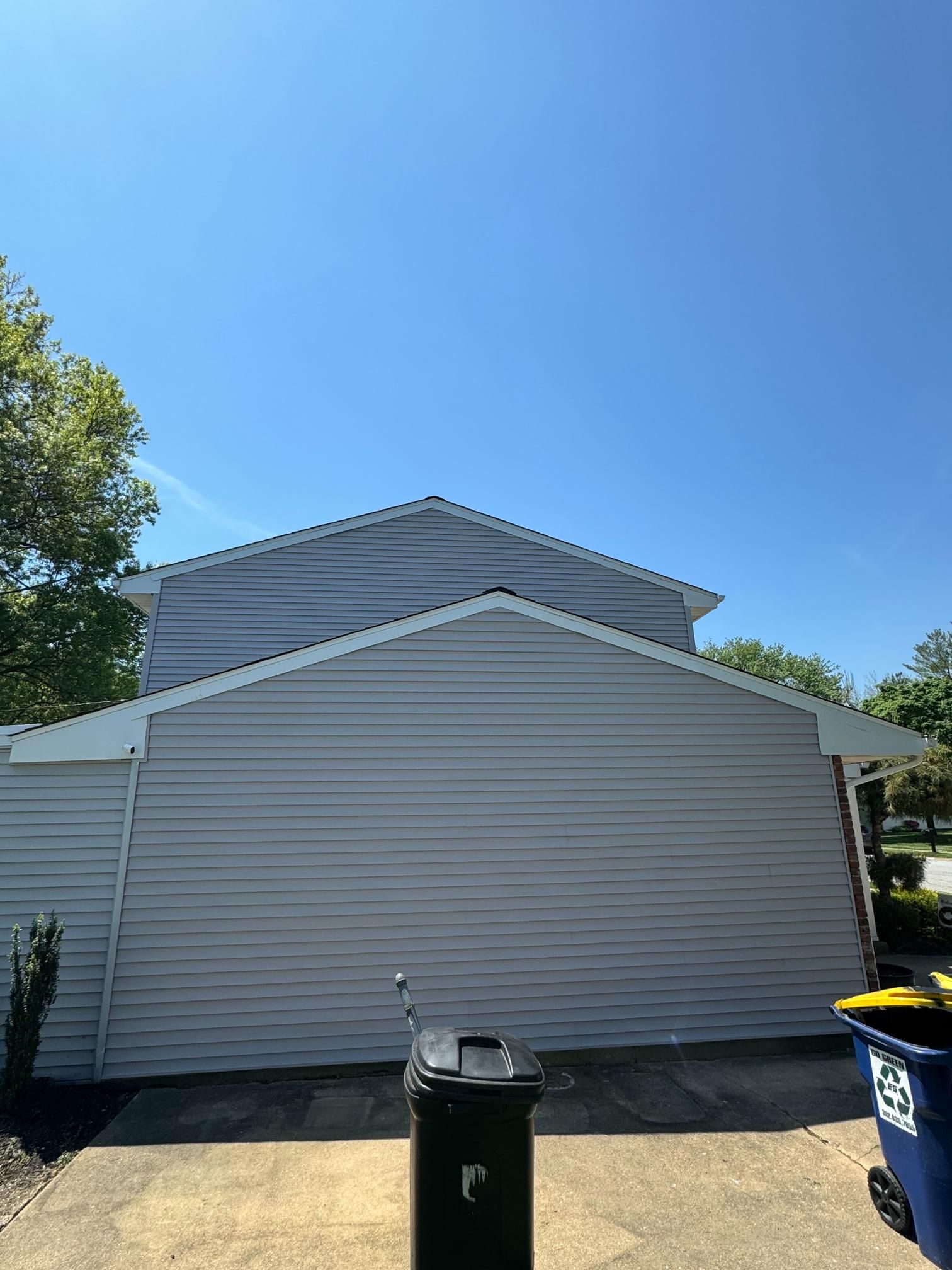 A two-story house with gray siding under a clear blue sky. A trash can sits in front.