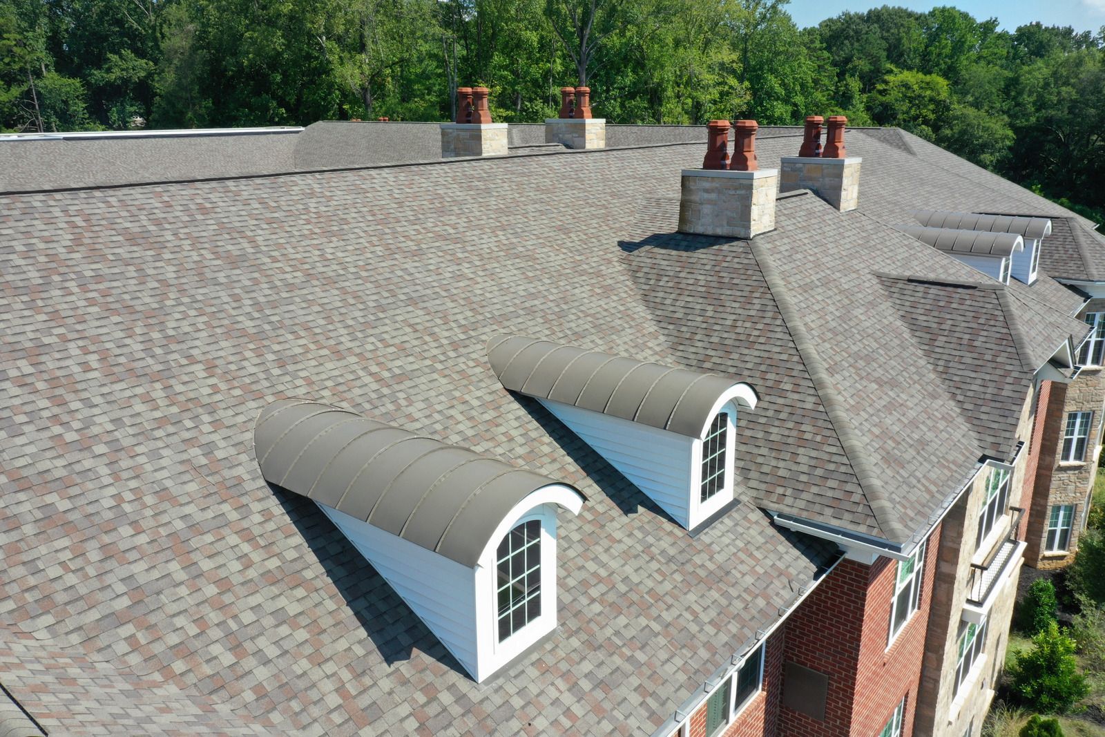 A building's roof with gray shingles, dormer windows, and brick chimneys against a tree-lined backdrop.