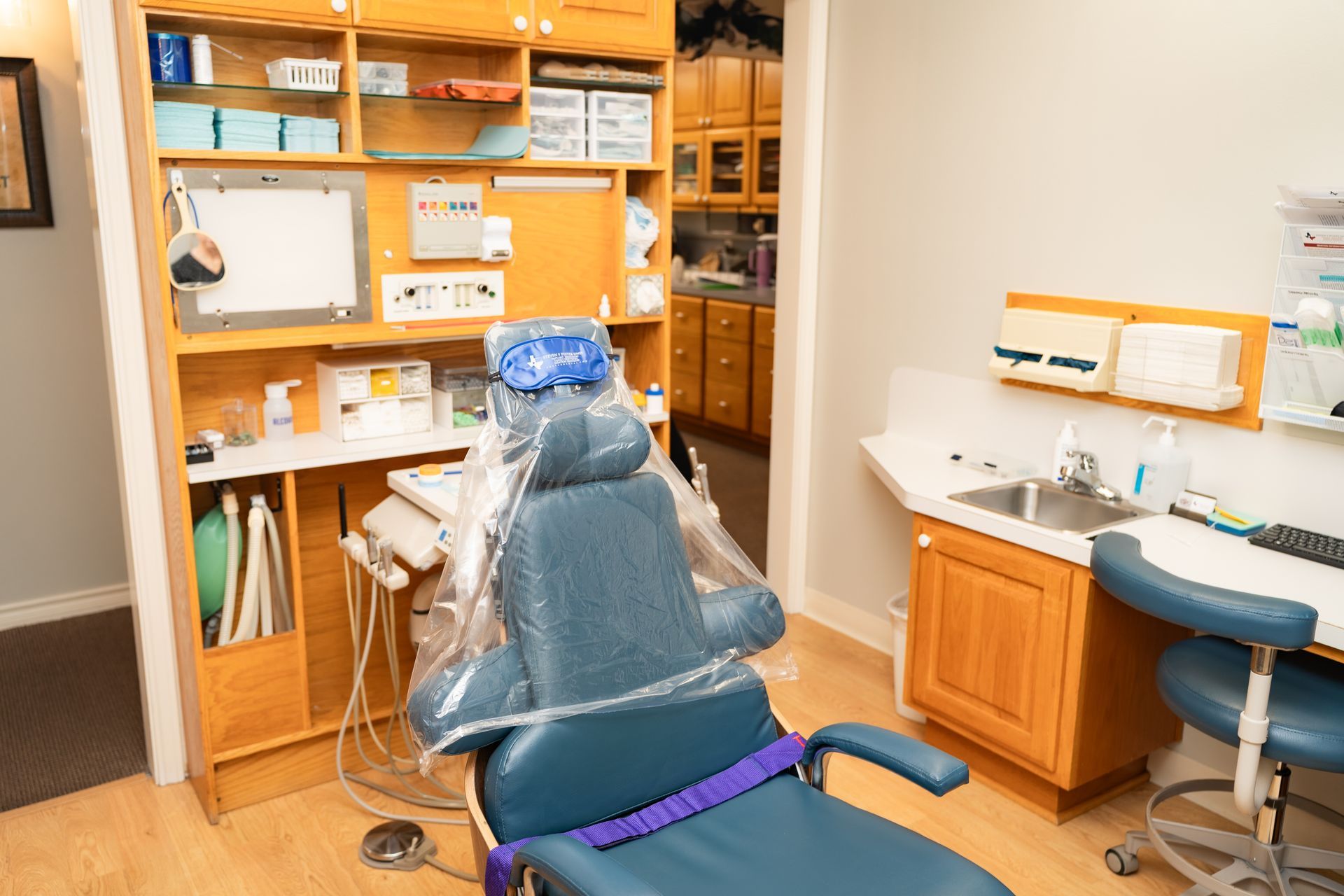 Dental examination room featuring a blue patient chair, wooden cabinets, a sink, and dental equipment.