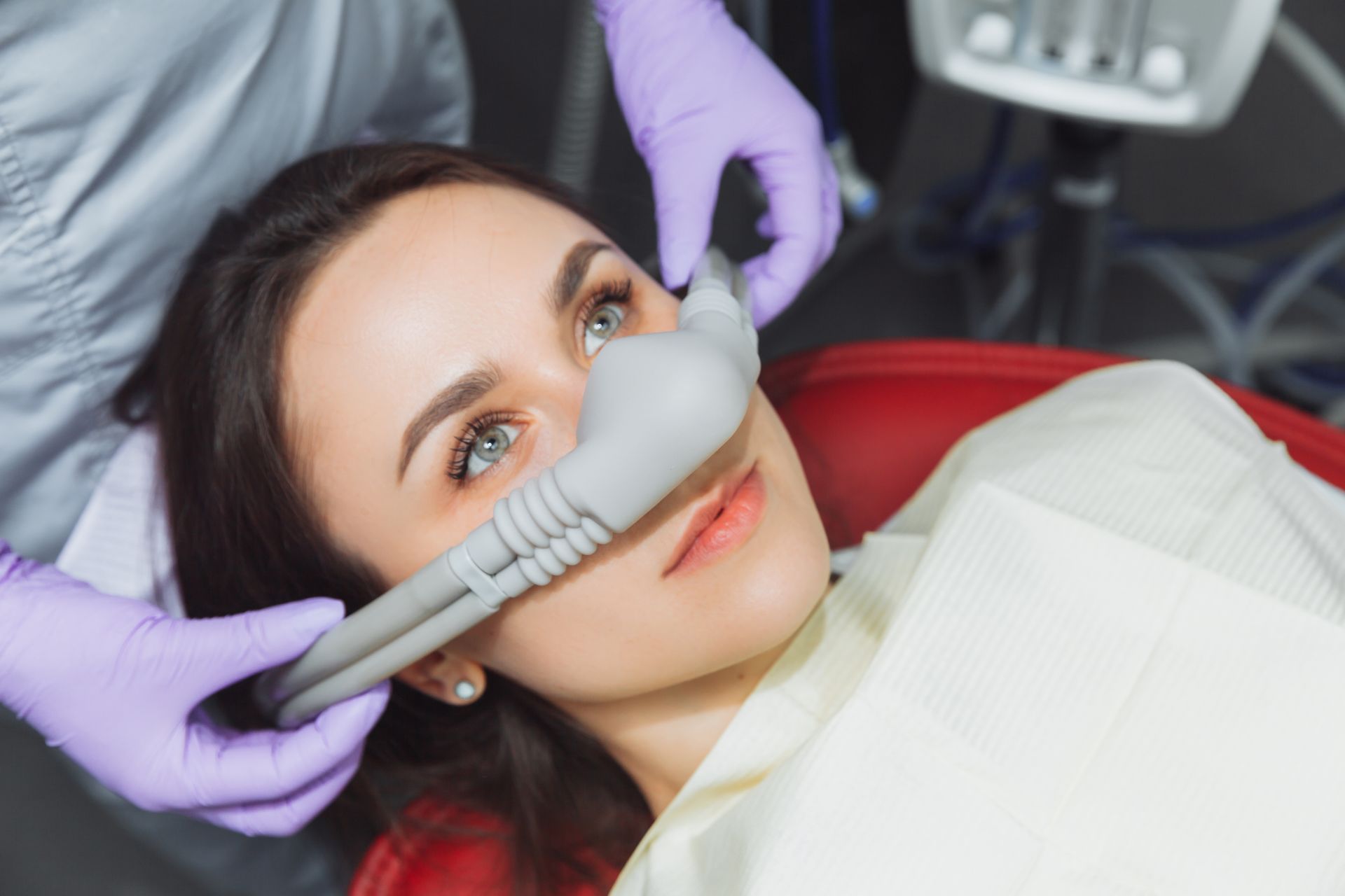 A patient in a dental chair wearing a gray nasal mask for sedation, with a practitioner's gloved hands holding it in place.
