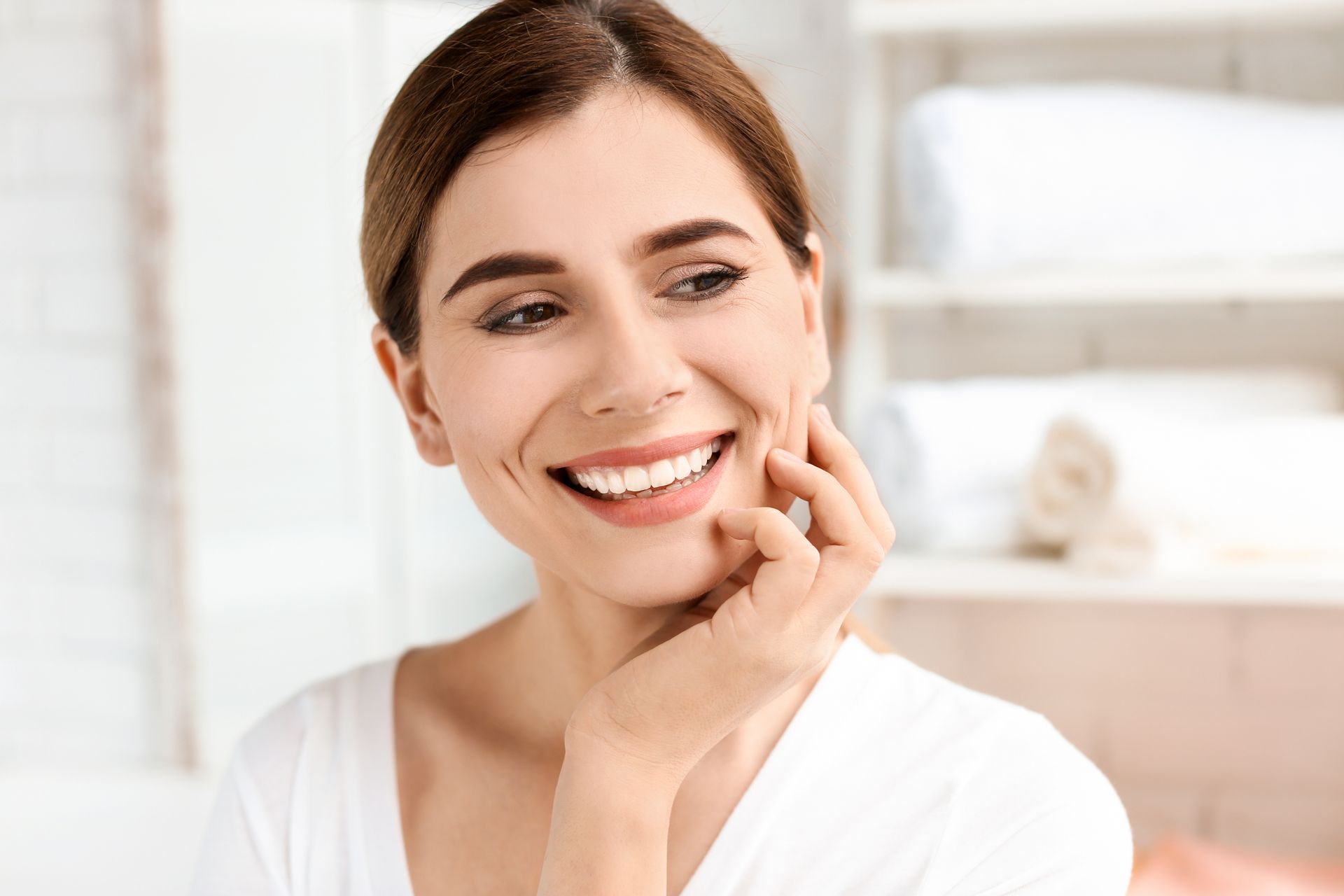 A person with a smiling expression touches their cheek, standing in a bright room with rolled white towels in the background.