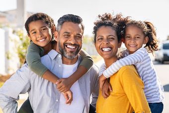 Two parents smiling while giving their two children piggyback rides outdoors on a sunny day.